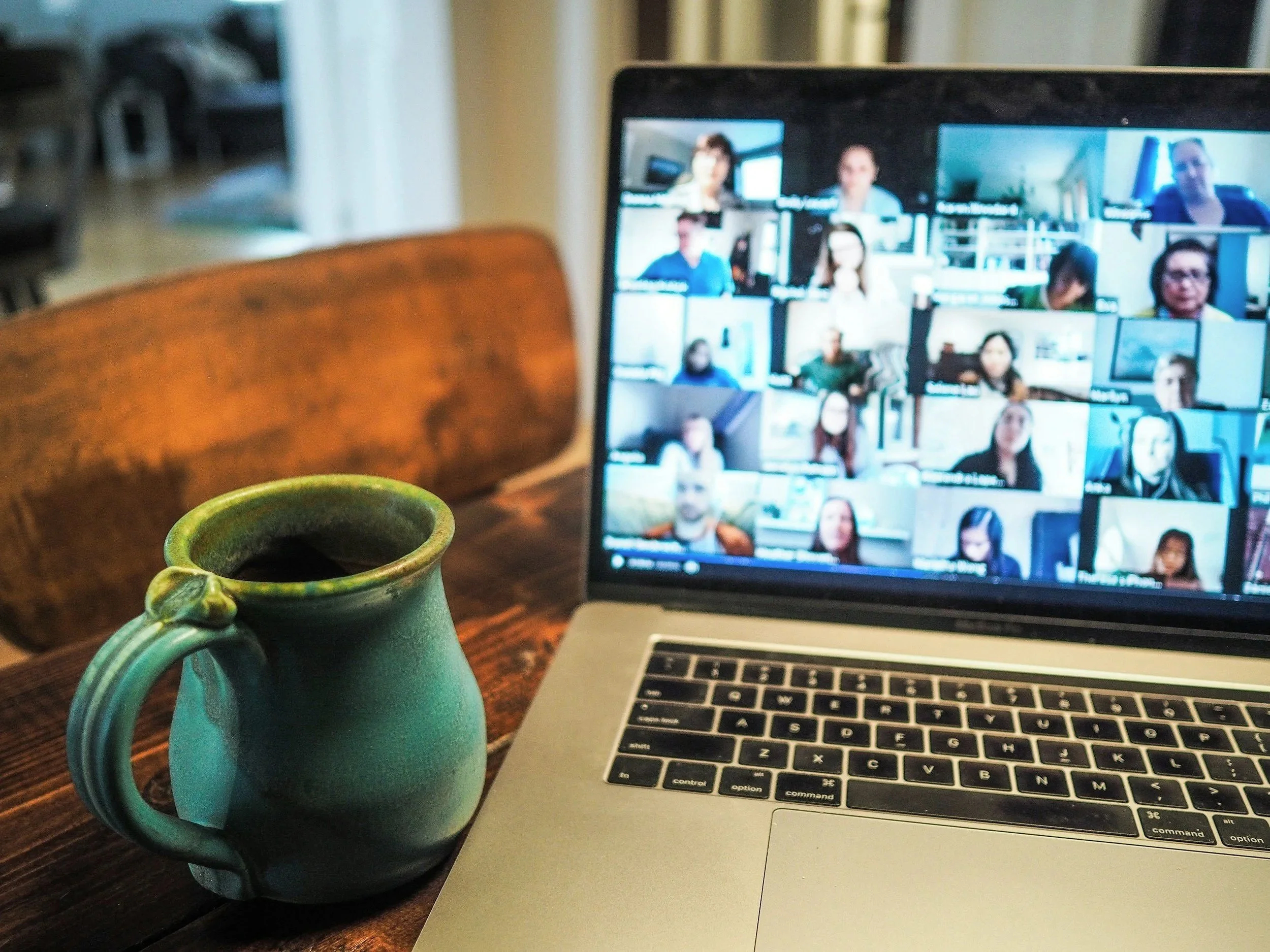 A laptop showing a virtual meeting with multiple participants on the screen, placed on a wooden table next to a green ceramic coffee mug.