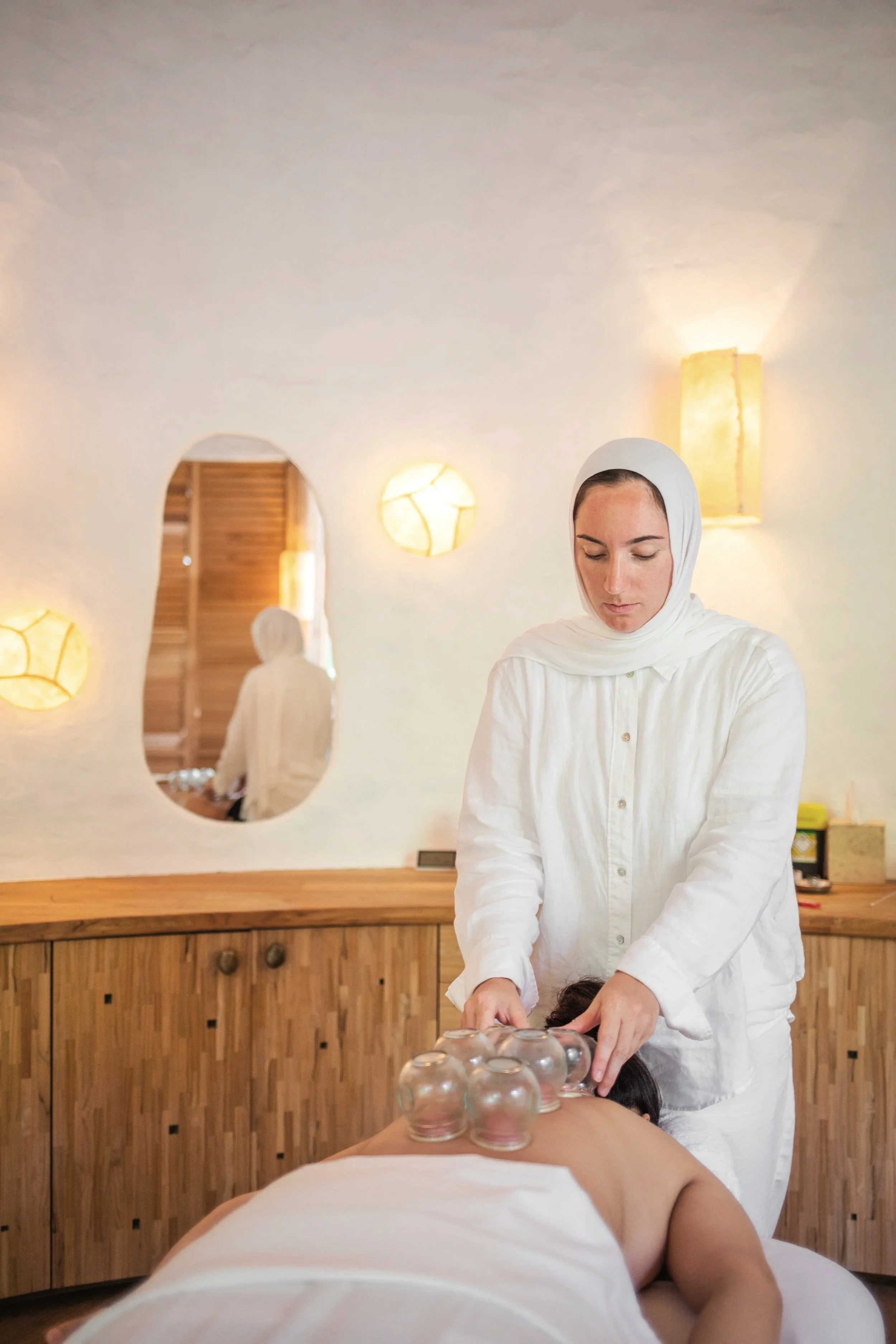 A woman receiving a glass cup therapy or massage in a spa-like setting, with another woman lying face down on a massage table, and soft lighting and decor in the background.