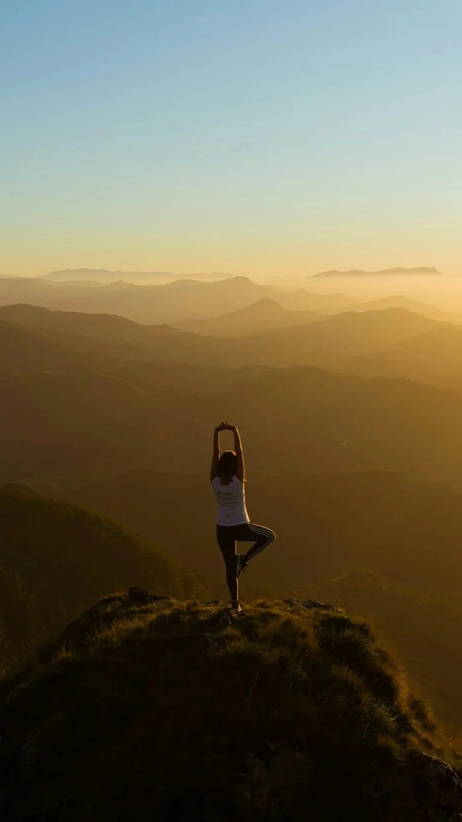 A person practicing yoga on top of a mountain at sunset, standing in a tree pose with arms raised overhead, overlooking a vast mountain range.