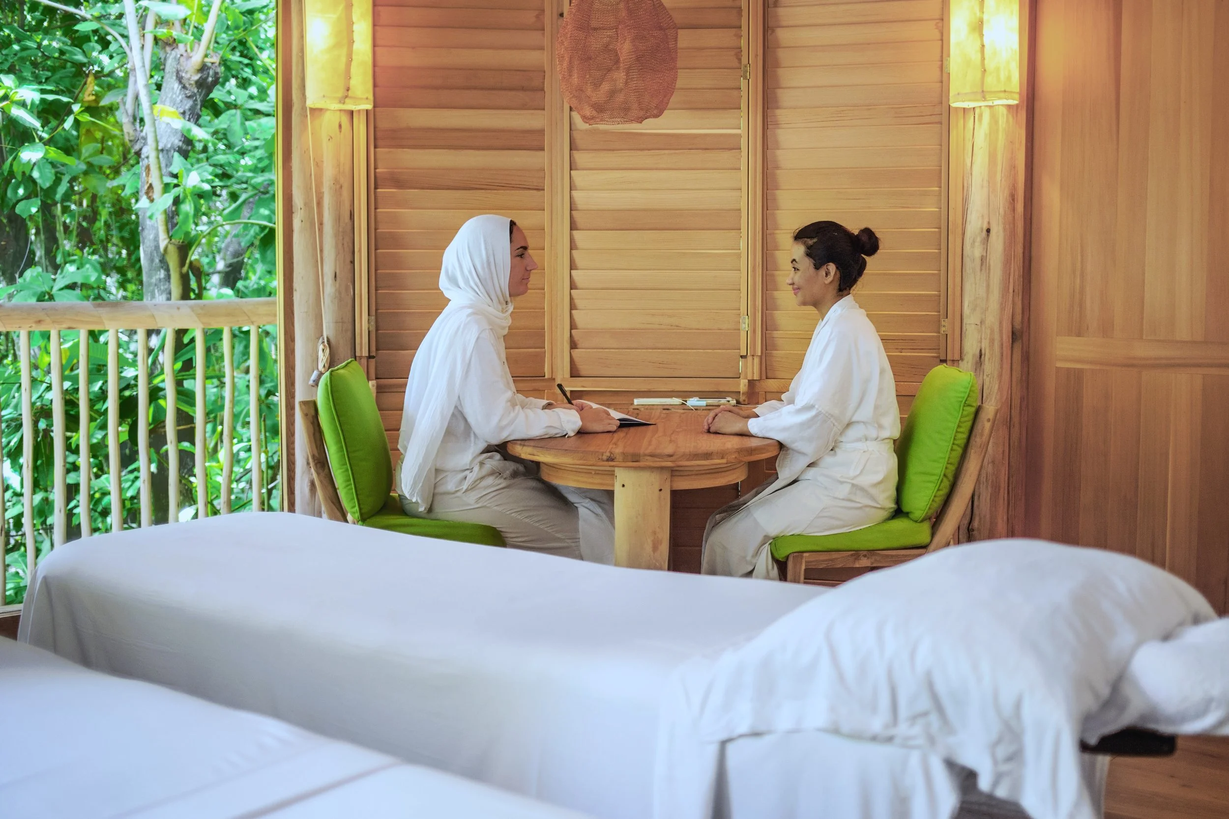 Two women sit at a wooden table with green cushions in a room with wooden walls and open window showing a lush green outdoor view. They are dressed in white, facing each other, engaged in a conversation.