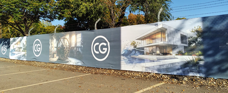 A construction fence banner depicting architectural designs of modern residential houses, placed beside an empty parking lot with trees and power lines in the background.
