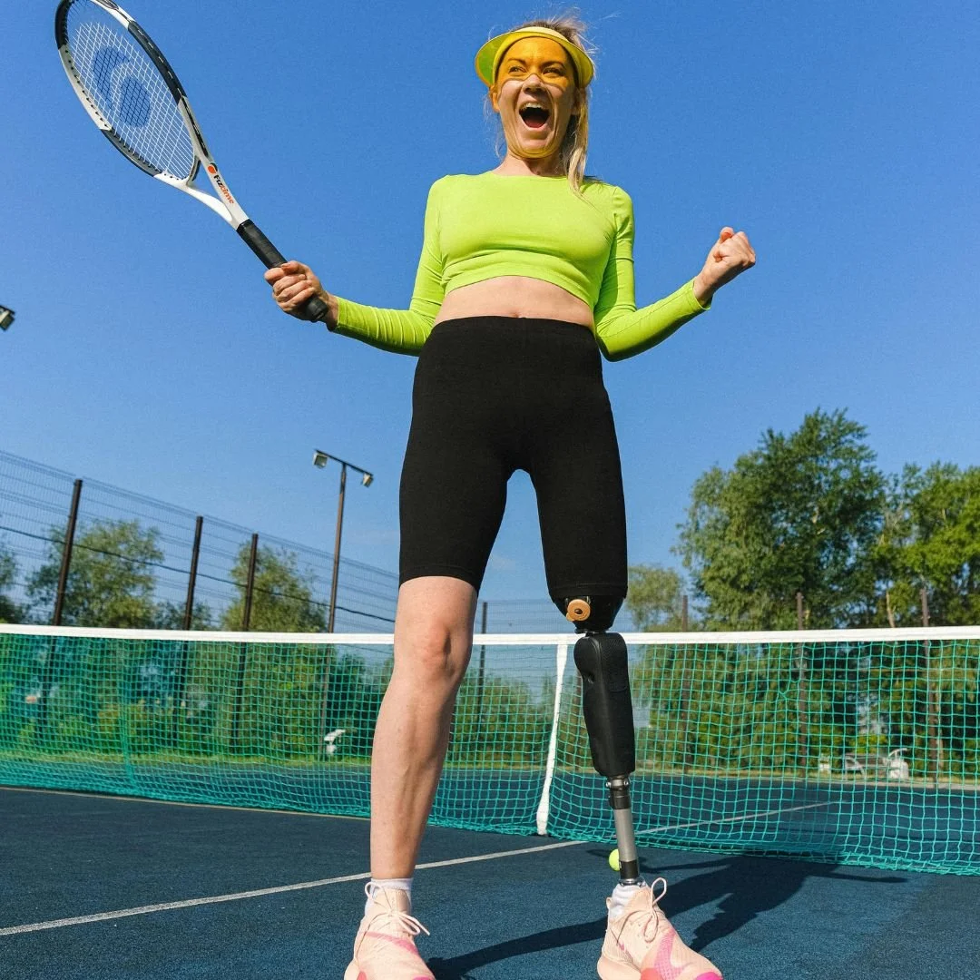 Joyful female tennis player with a prosthetic leg celebrating a point on an outdoor tennis court, holding a racket and wearing a bright green long-sleeve crop top and black shorts.