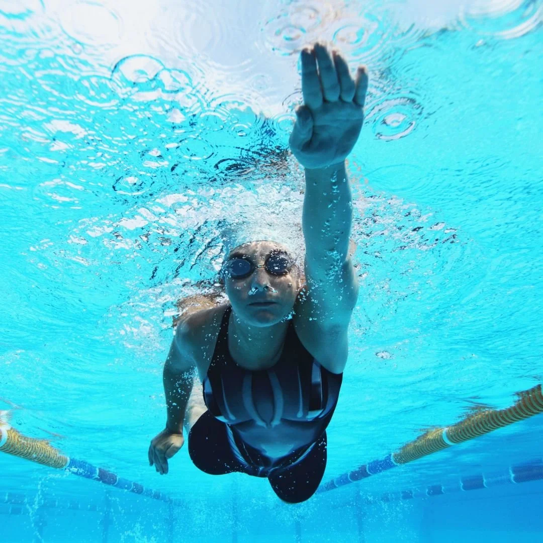 An underwater view of a female swimmer in a black swimsuit, goggles, and swim cap performing the front crawl stroke in a pool.