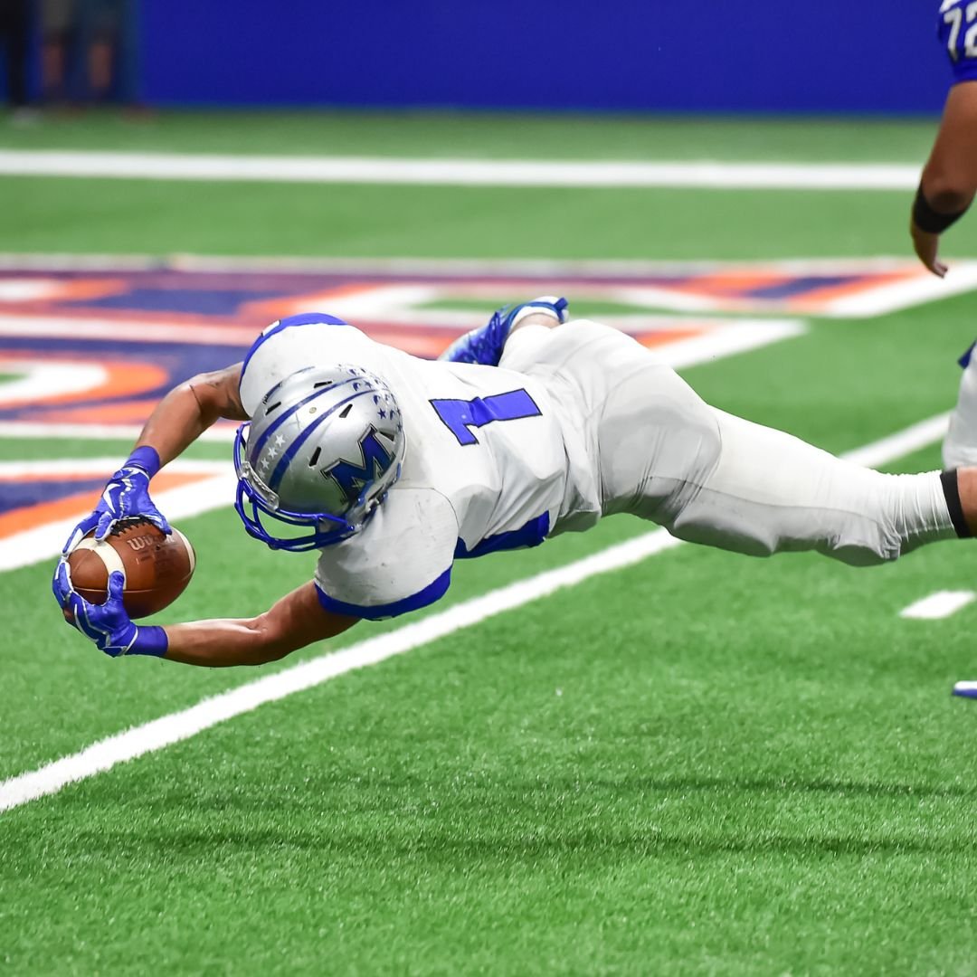 American football player in a white uniform diving horizontally to catch the football on the turf.