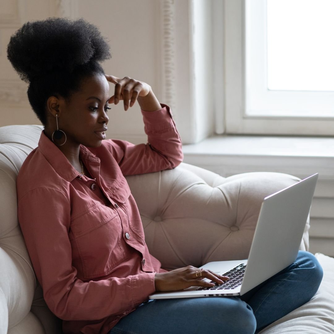 Young Black woman sitting cross-legged on a cream armchair working thoughtfully on a laptop.