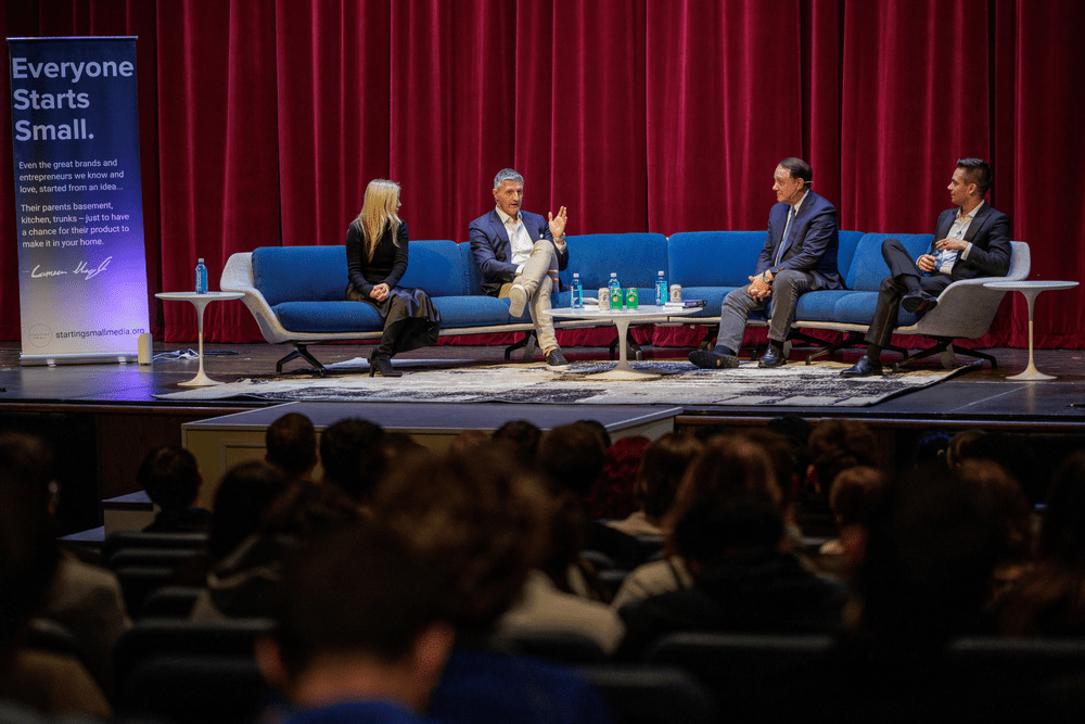 Panelists on a blue sofa in front of a full auditorium in 2025