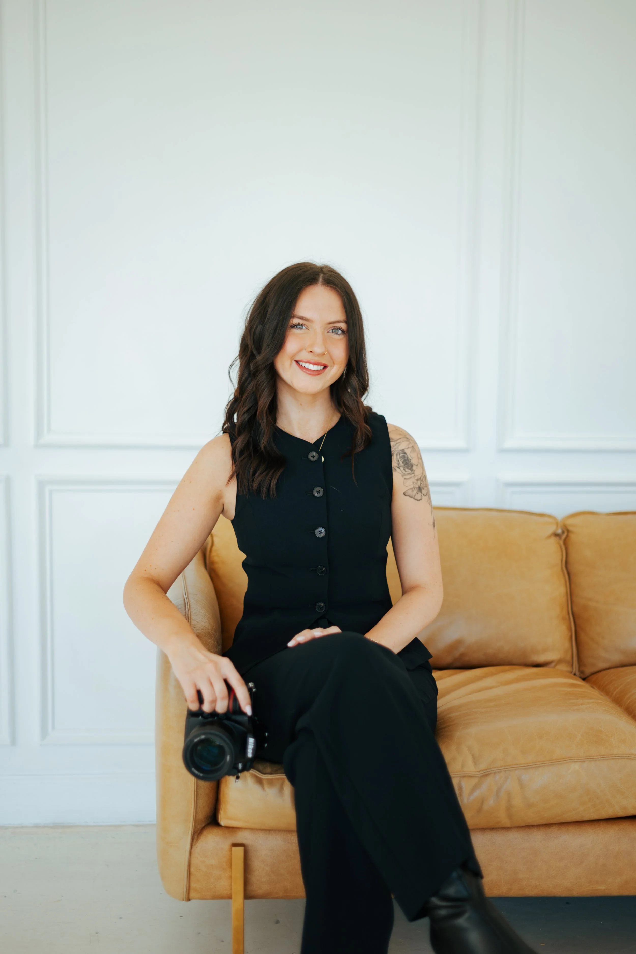 A woman with shoulder-length dark hair, wearing a black sleeveless button-up top and black pants, sitting on a tan leather sofa in a white room, holding a professional camera in her right hand, smiling at the camera.