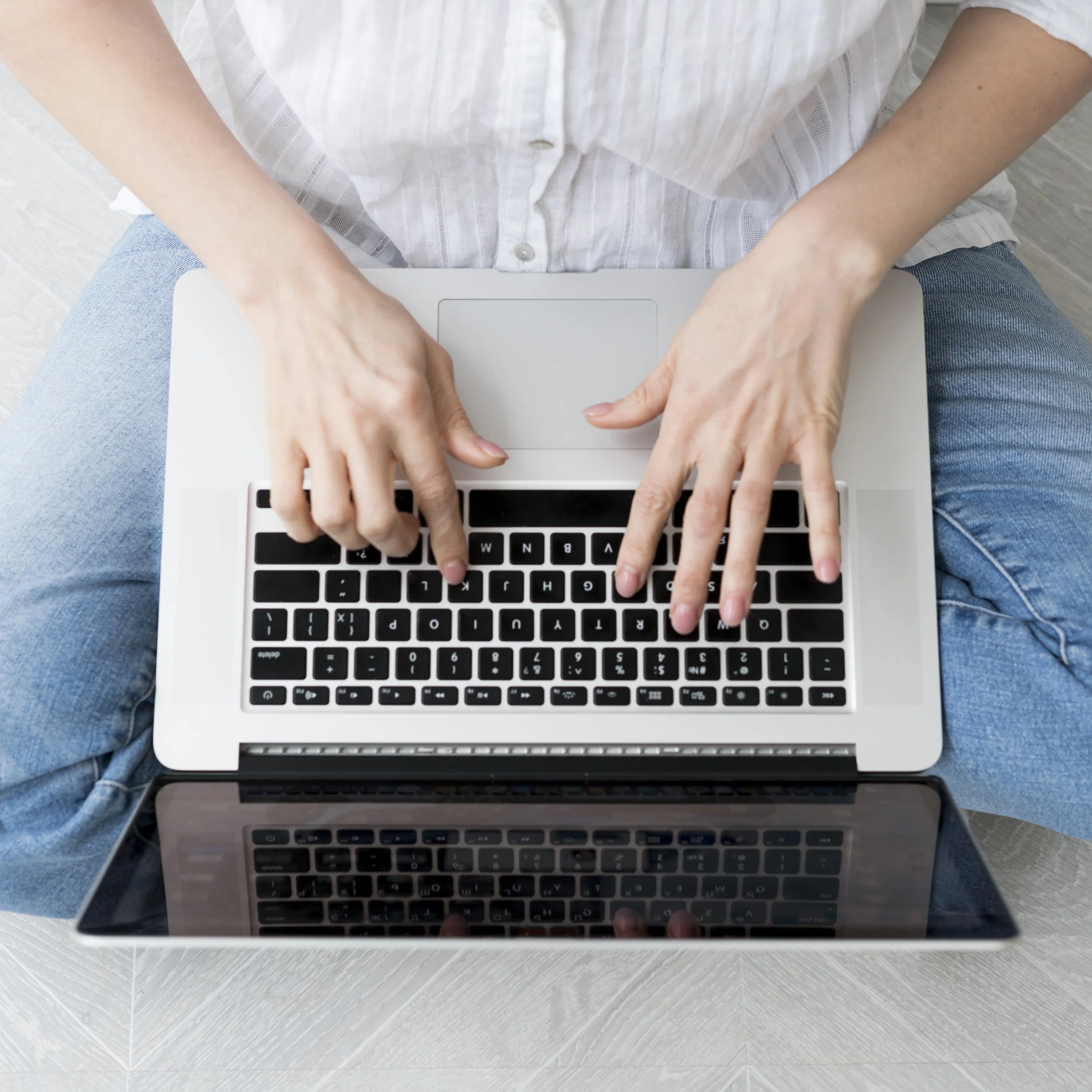 Person sitting cross-legged on a light-colored wooden floor, using a silver laptop with a black keyboard.