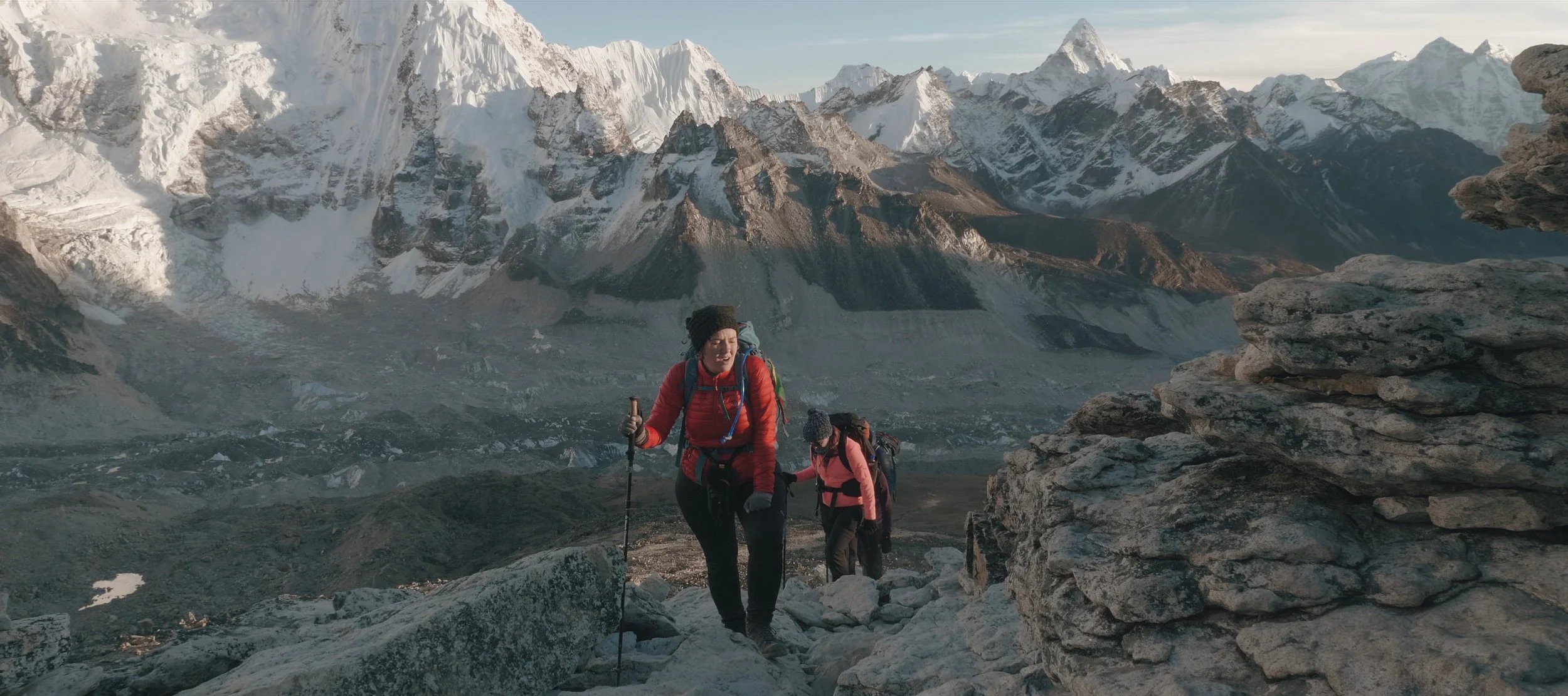 Two hikers trekking in a mountainous, snow-covered landscape with rocky terrain and towering peaks in the background.