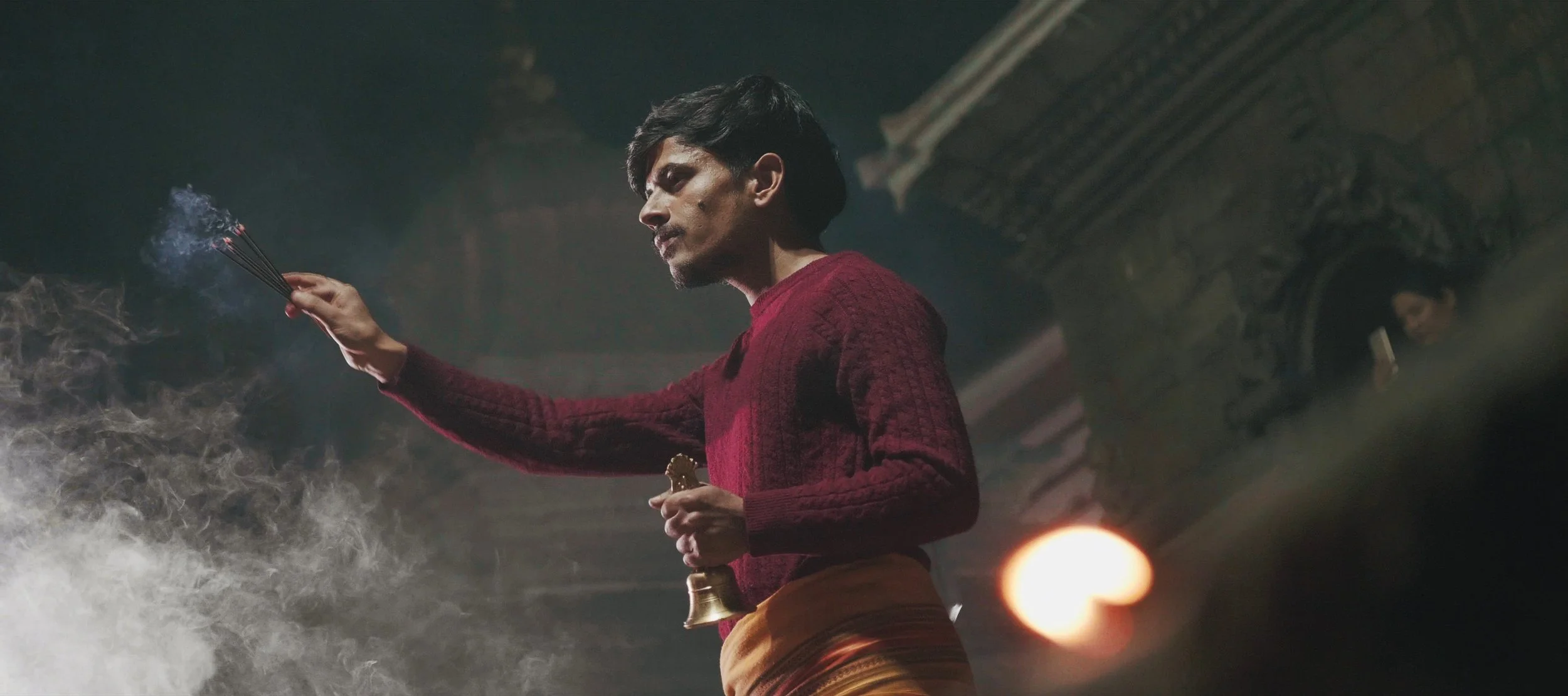A man in a red sweater holding a bundle of lit incense sticks in one hand and a small brass bell in the other, standing in a dimly lit indoor setting with ornate architectural details.