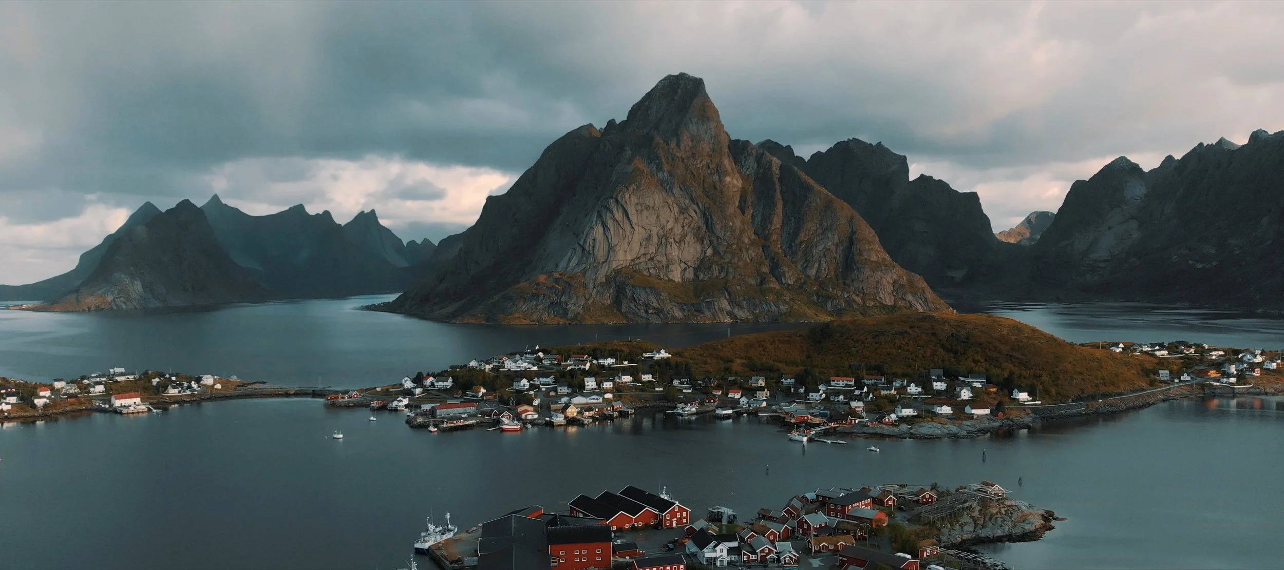 Scenic view of a small village with colorful houses on an island, surrounded by a calm body of water and backed by towering, rugged mountains under a cloudy sky.