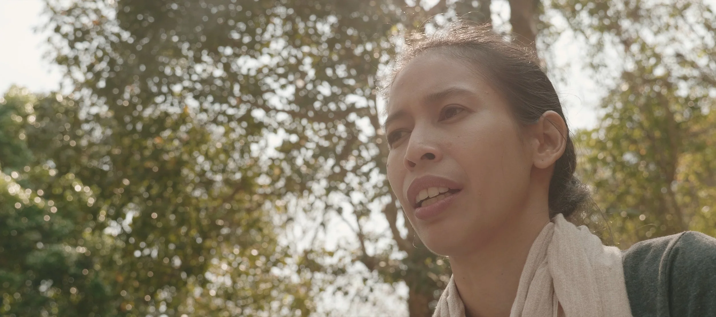 Close-up of a woman outdoors with trees and sunlight in the background.