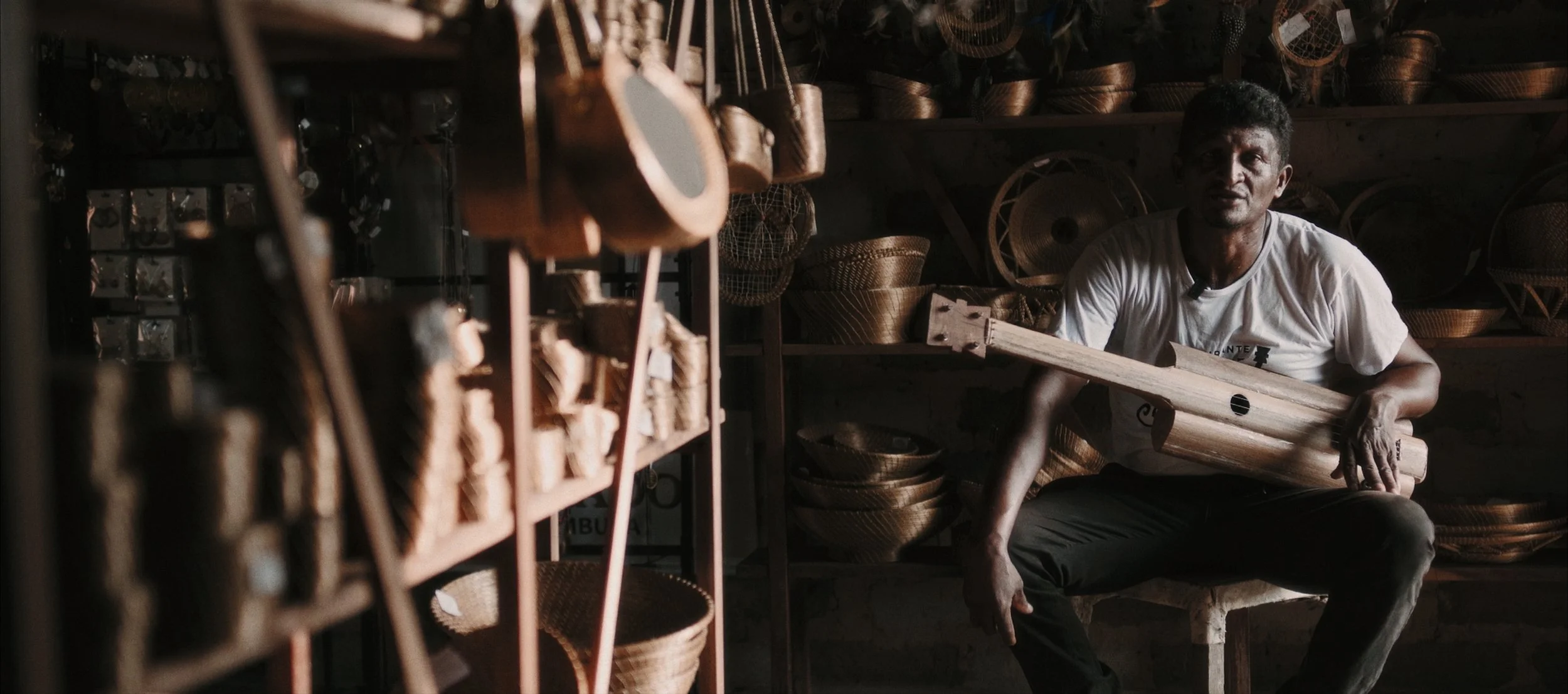 Man sitting in a basket shop holding a woven basket with shelves of baskets around him.