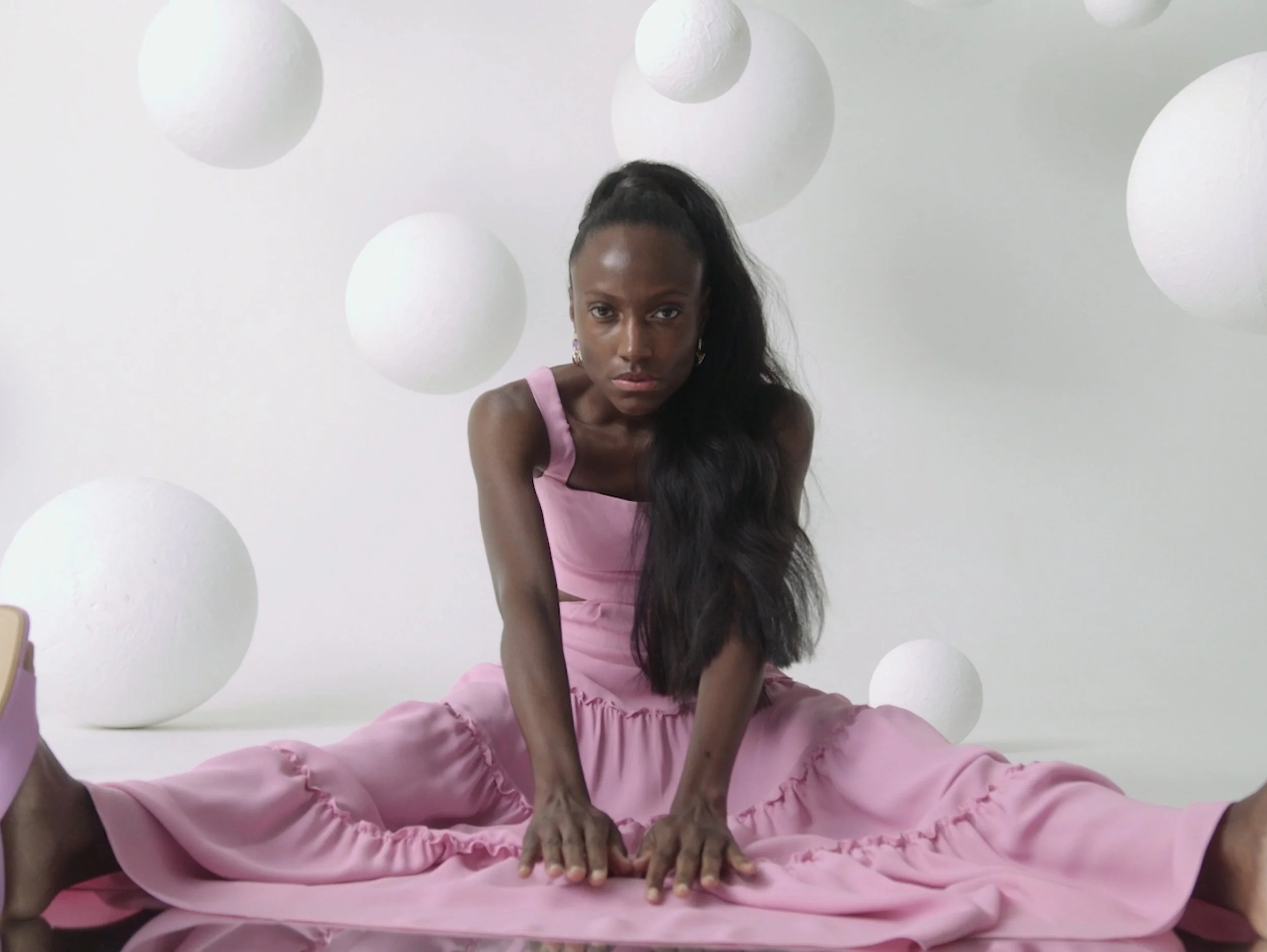 A woman in a pink dress sitting cross-legged on the floor, surrounded by white spherical objects, against a white background.