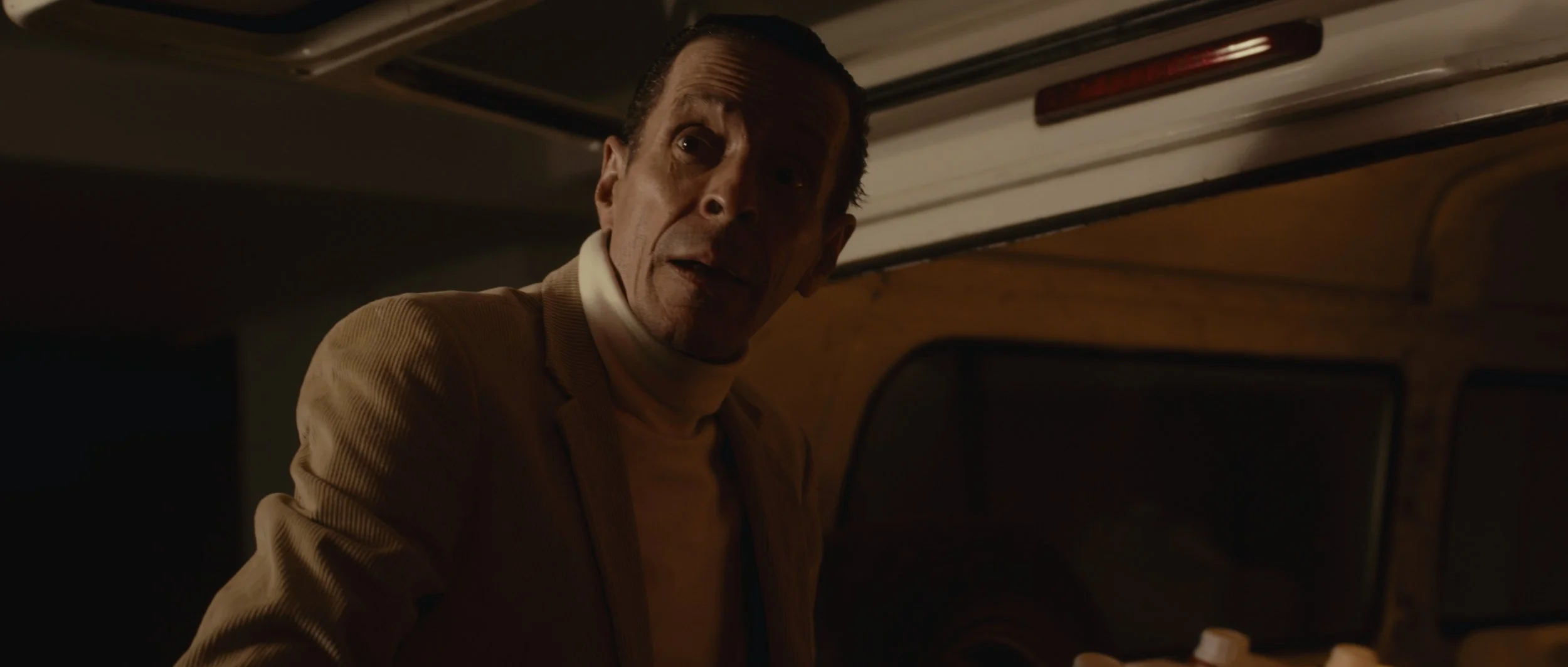 A man with slicked-back hair and wearing a beige jacket and white turtleneck looking over his shoulder in a dimly lit setting.