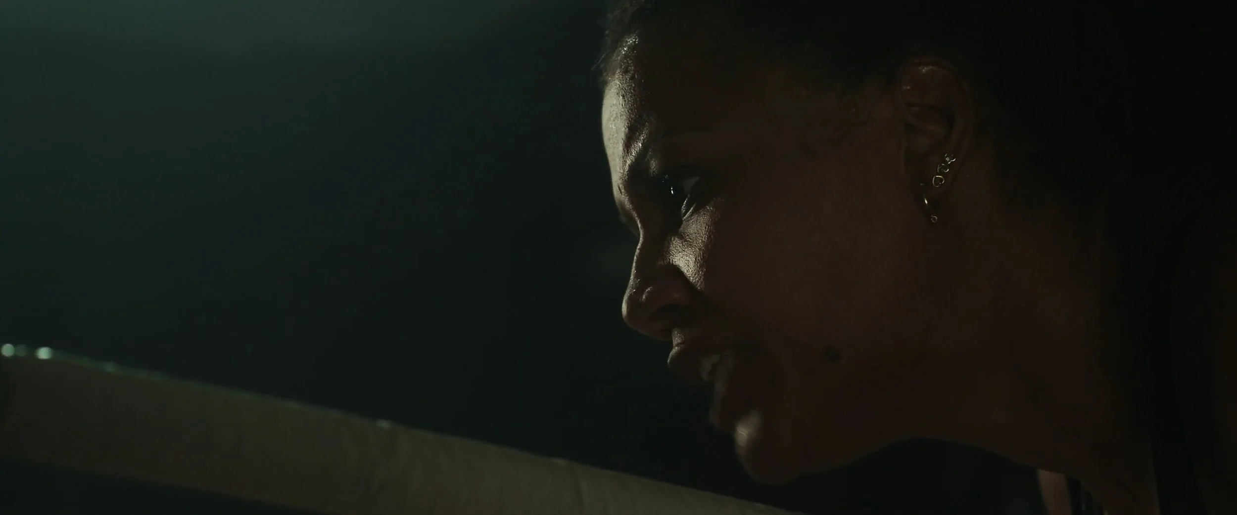 Close-up of a woman with earrings, appears to be in a dimly lit setting, looking downward with a serious expression.