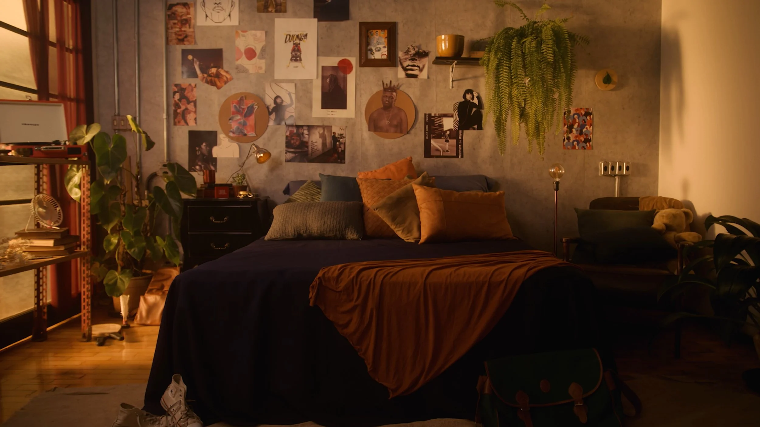 Cozy bedroom with a wall decorated with various art prints and photographs, a bed with orange and dark-colored pillows and a throw blanket, plants, a bookshelf, a nightstand, and a soft chair with plush toys in a warmly lit room.