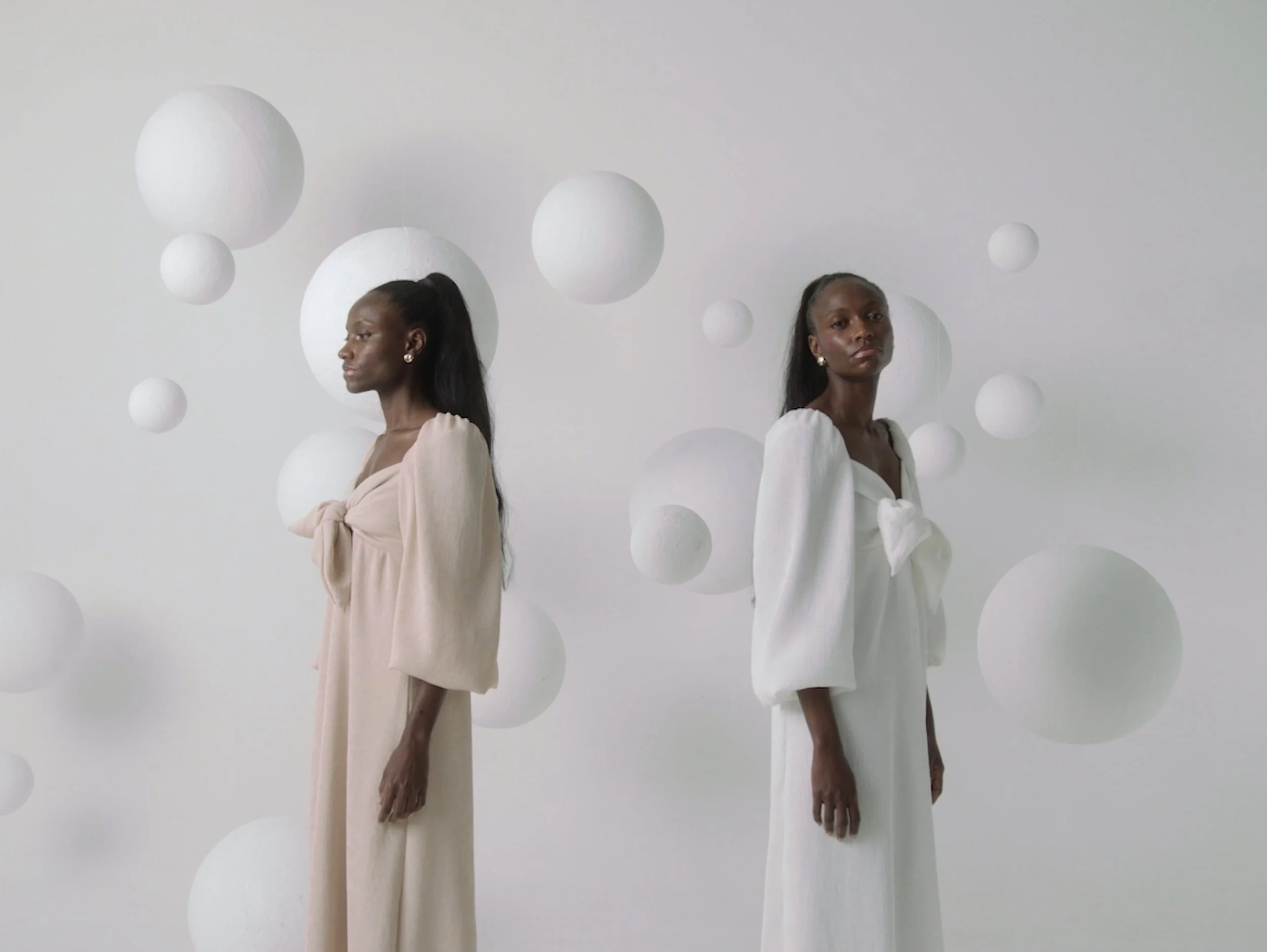 Two women with dark skin and long black hair standing side by side against a white background with floating white spheres. One woman is dressed in a light beige gown with a bow on the chest, and the other in an off-white dress with a bow on the front