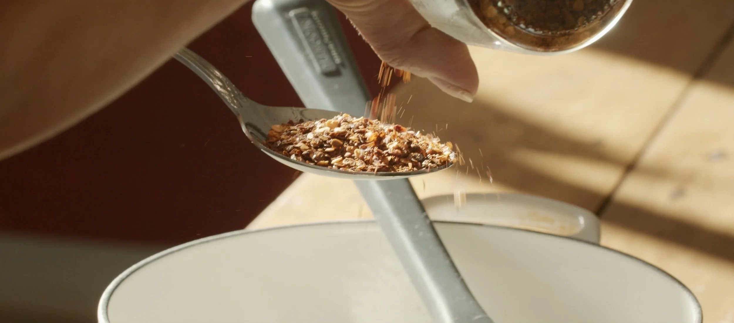 A hand sprinkling granola into a bowl from a spoon.