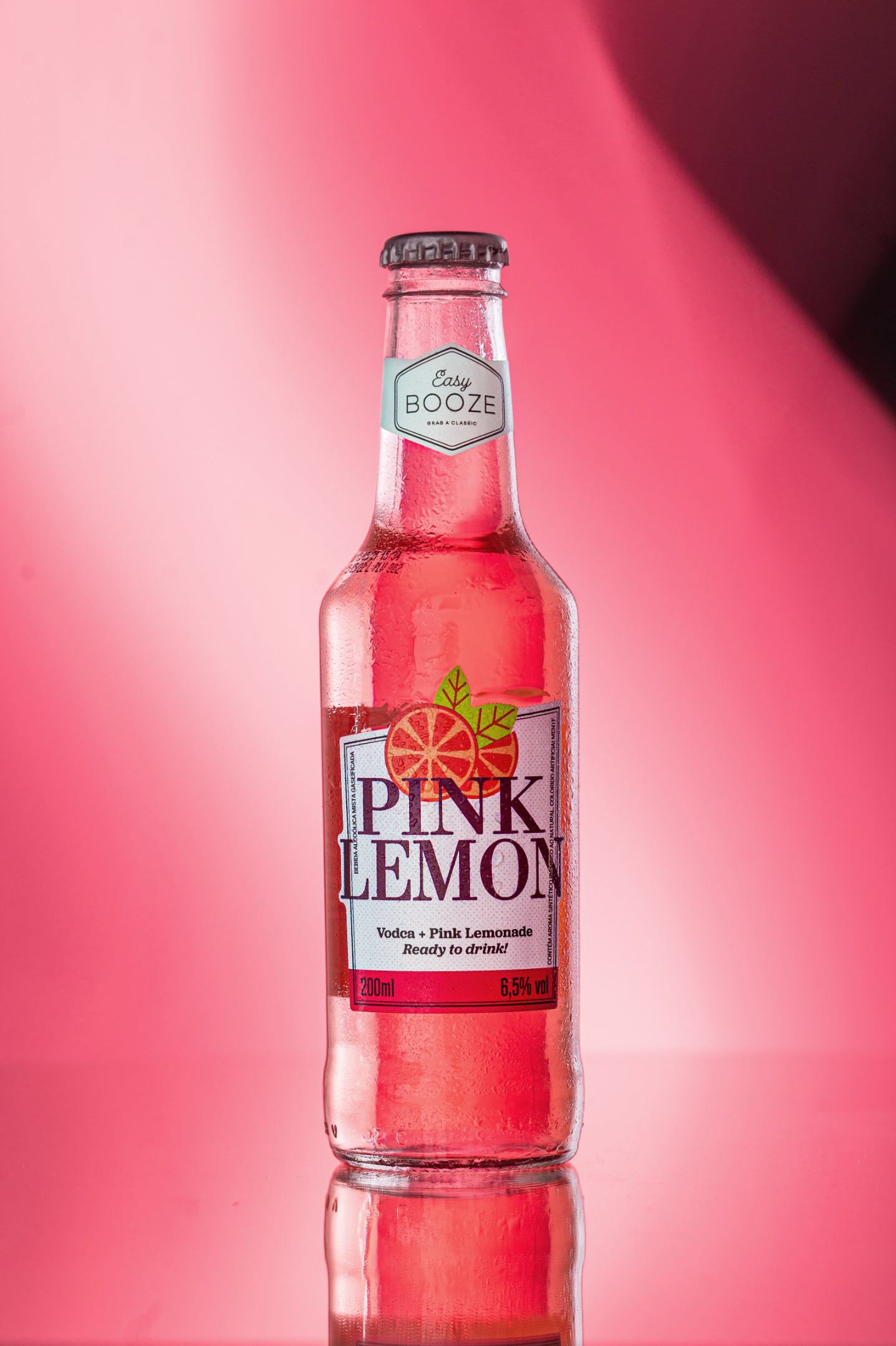 A pink lemonade flavored beverage in a glass bottle with a black cap, set against a pink background. The label on the bottle reads 'Pink Lemon' and indicates it contains vodka and pink lemonade, with 6.5% alcohol by volume.