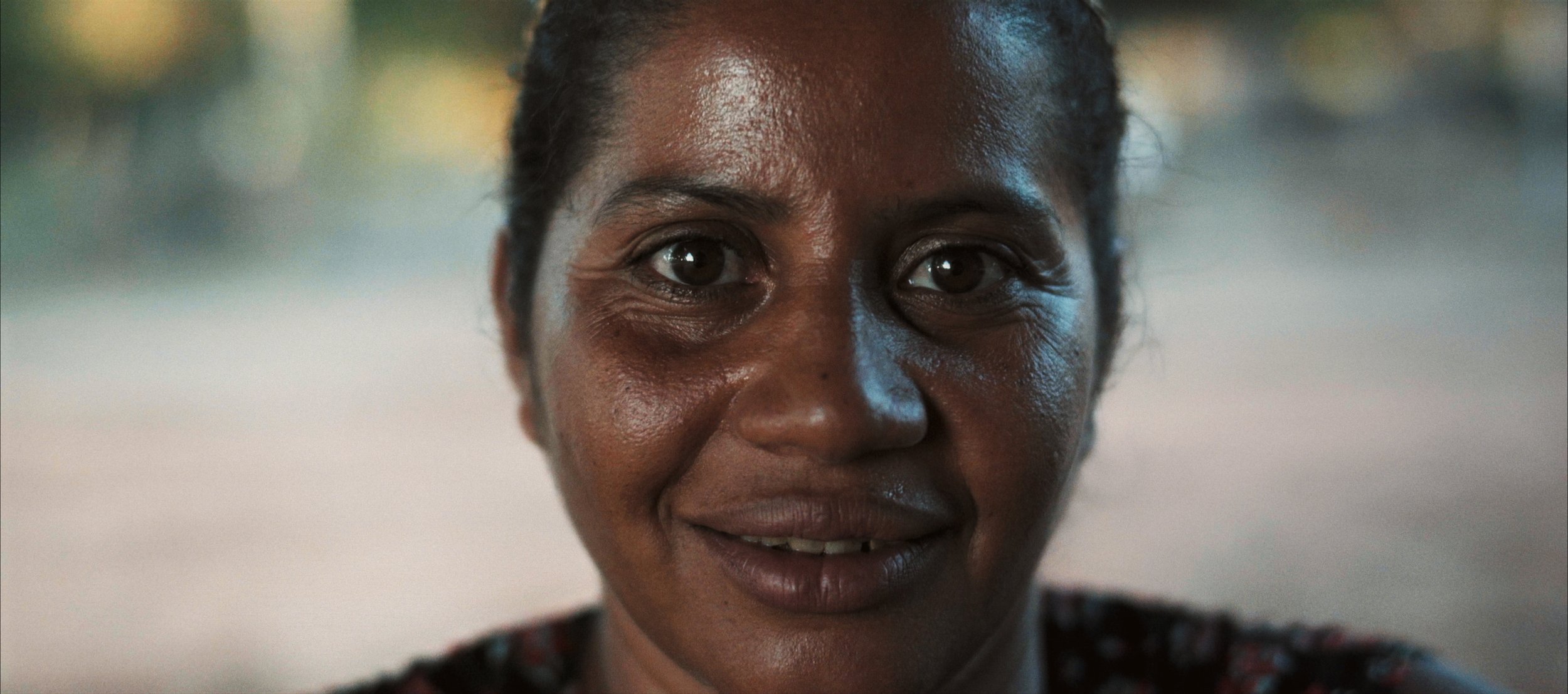 Close-up of a smiling woman with dark skin and dark hair, outdoors, with blurred background.