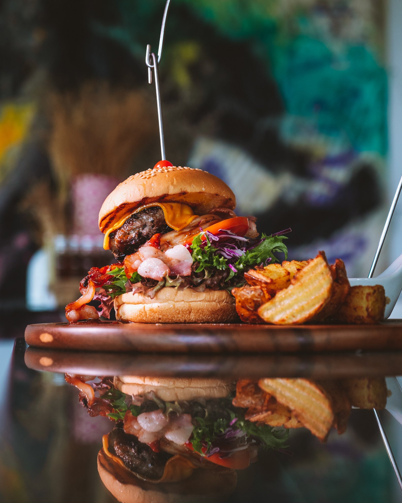 Close-up of a gourmet cheeseburger with bacon, lettuce, tomato, and cheese, served with potato wedges on a wooden plate with a colorful background.