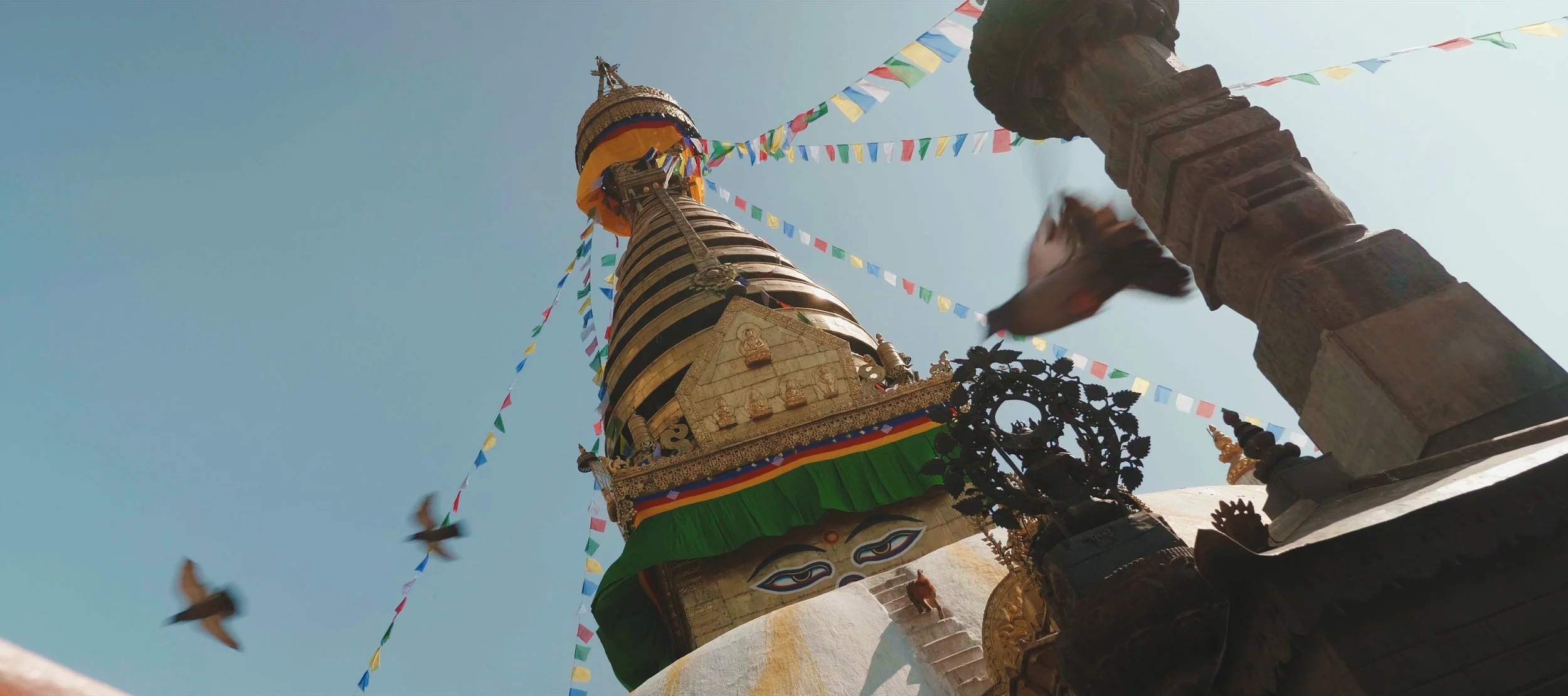 A towering Buddhist stupa decorated with colorful prayer flags and intricate carvings, with a bright blue sky in the background.