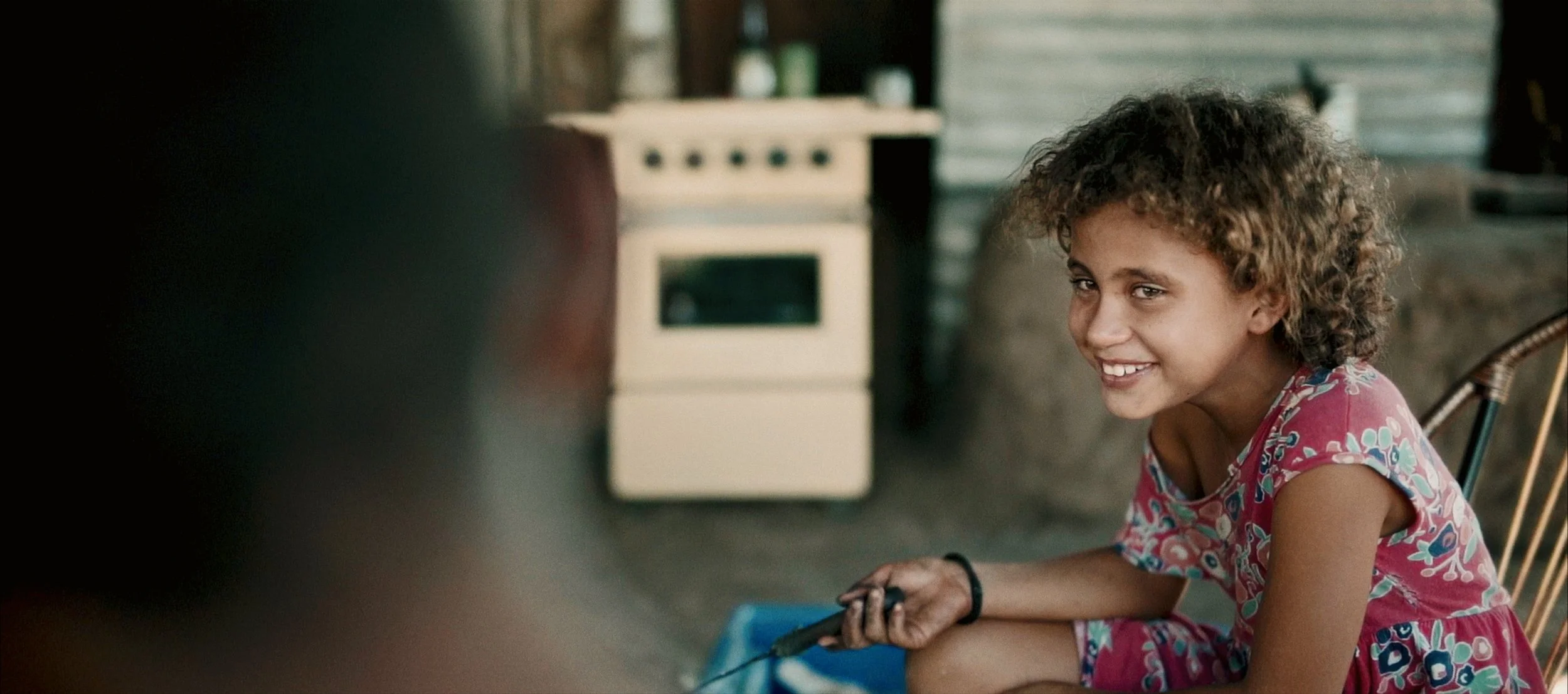 A young girl with curly hair smiling while holding a cleaning brush, sitting on a chair in a rustic kitchen with a stove in the background.