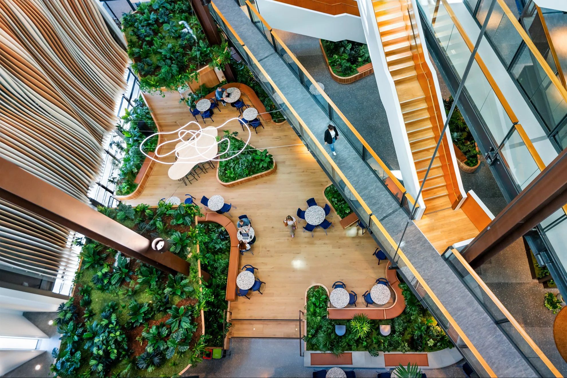 Interior view of a modern, multi-level building with a large open atrium, green plants, and seating areas with blue chairs and round tables. A few people are walking and sitting within the space, which features wooden flooring, glass walls, and a sta