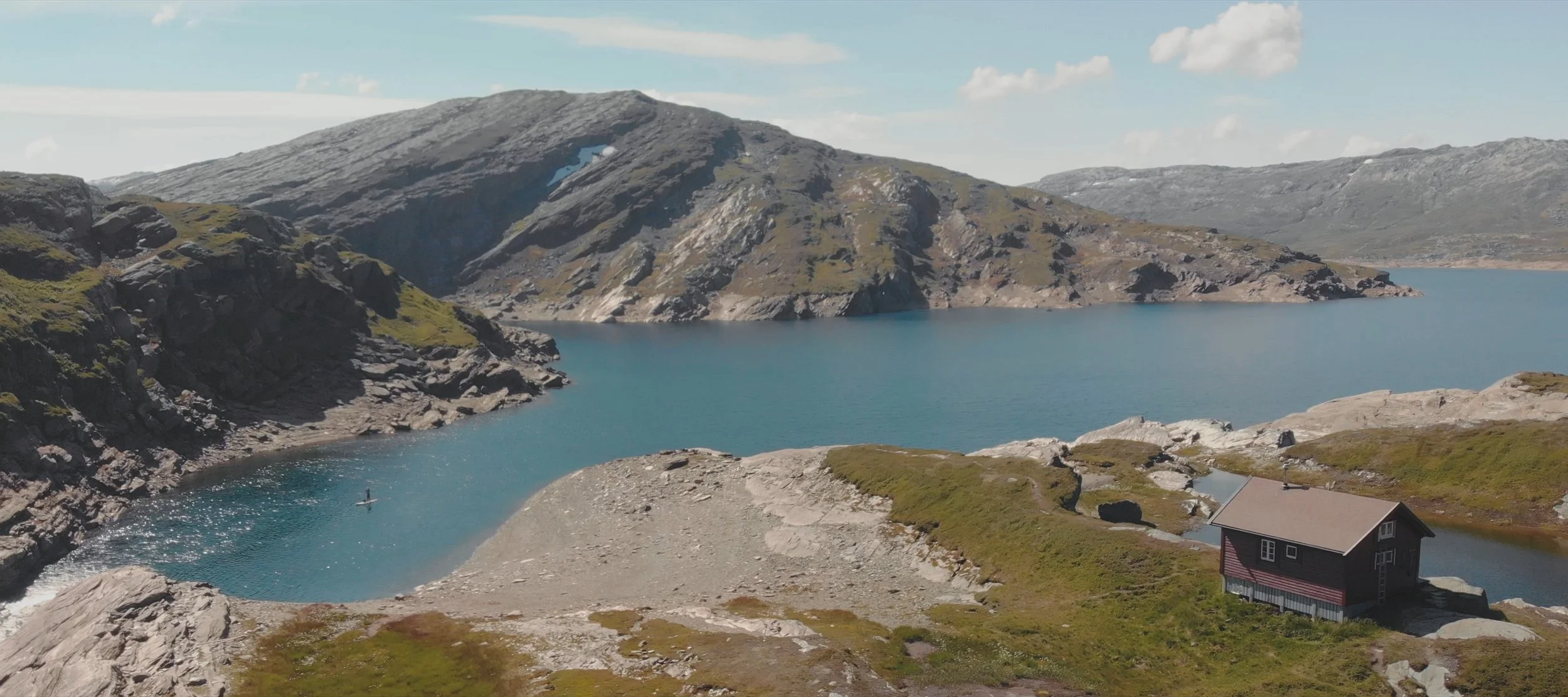 A mountain landscape with a large blue lake, rocky slopes, and a small black house with a brown roof near the water's edge.