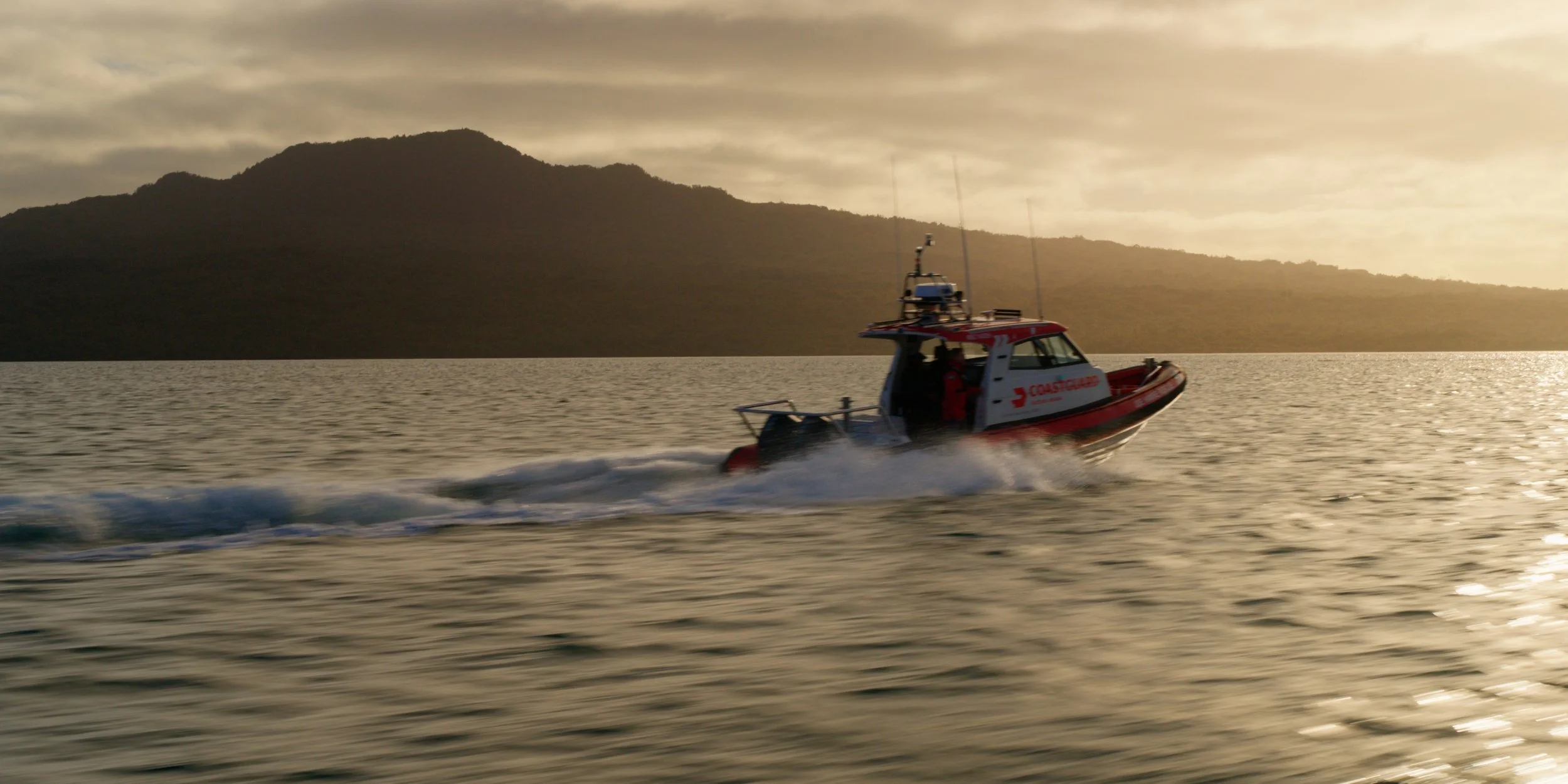 A Coast Guard rescue boat moving quickly across the water during sunset with mountains in the background.