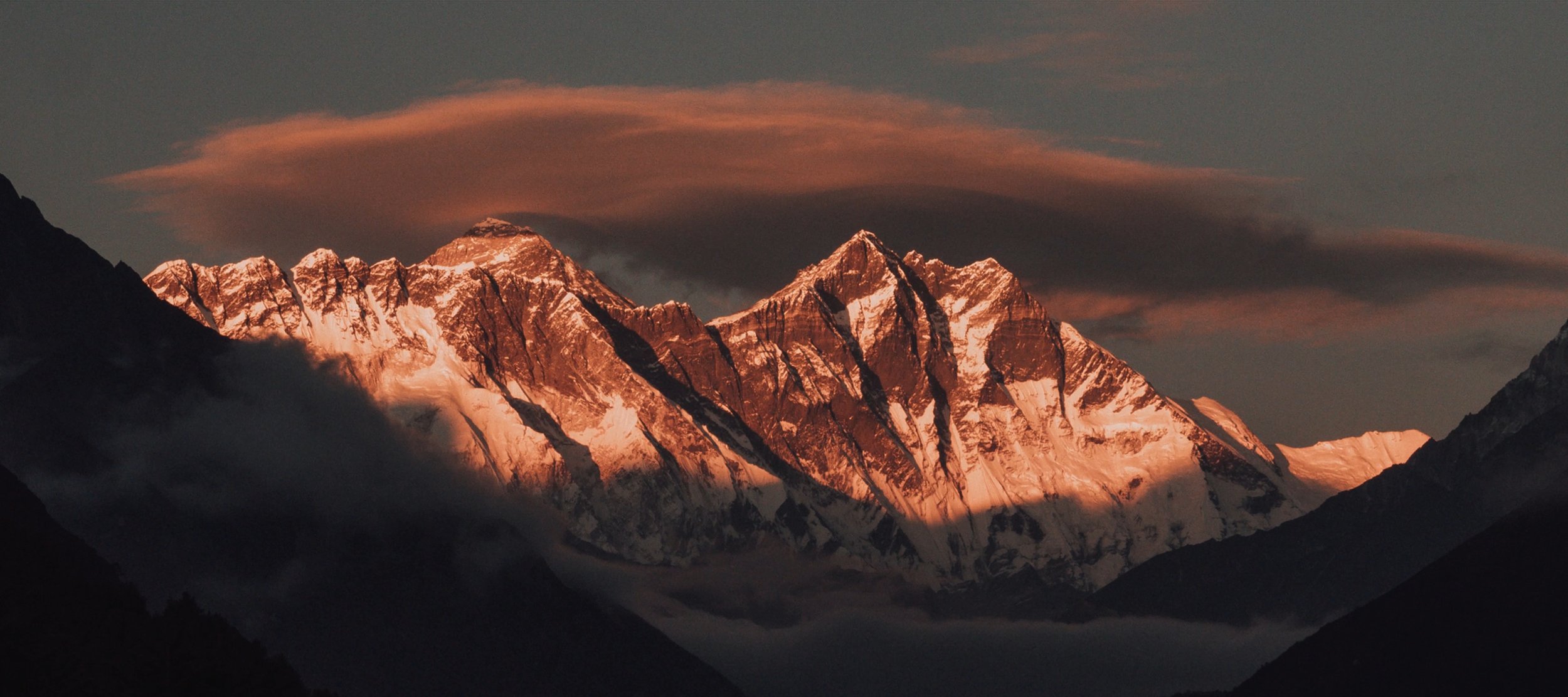 Snow-capped mountain peaks at sunset, with clouds and a darkened foreground.