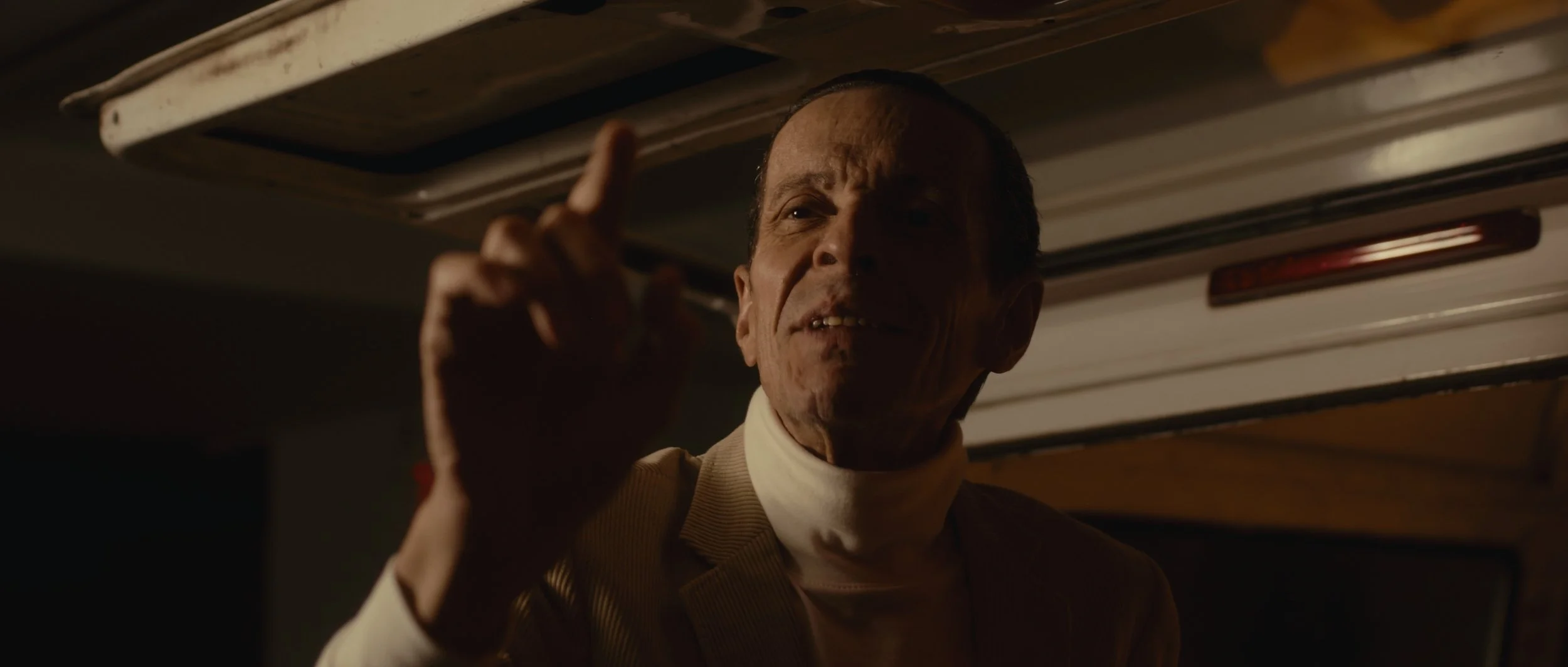 A man with slicked-back hair and a beige jacket, pointing with his index finger, standing under a ceiling with a light fixture.
