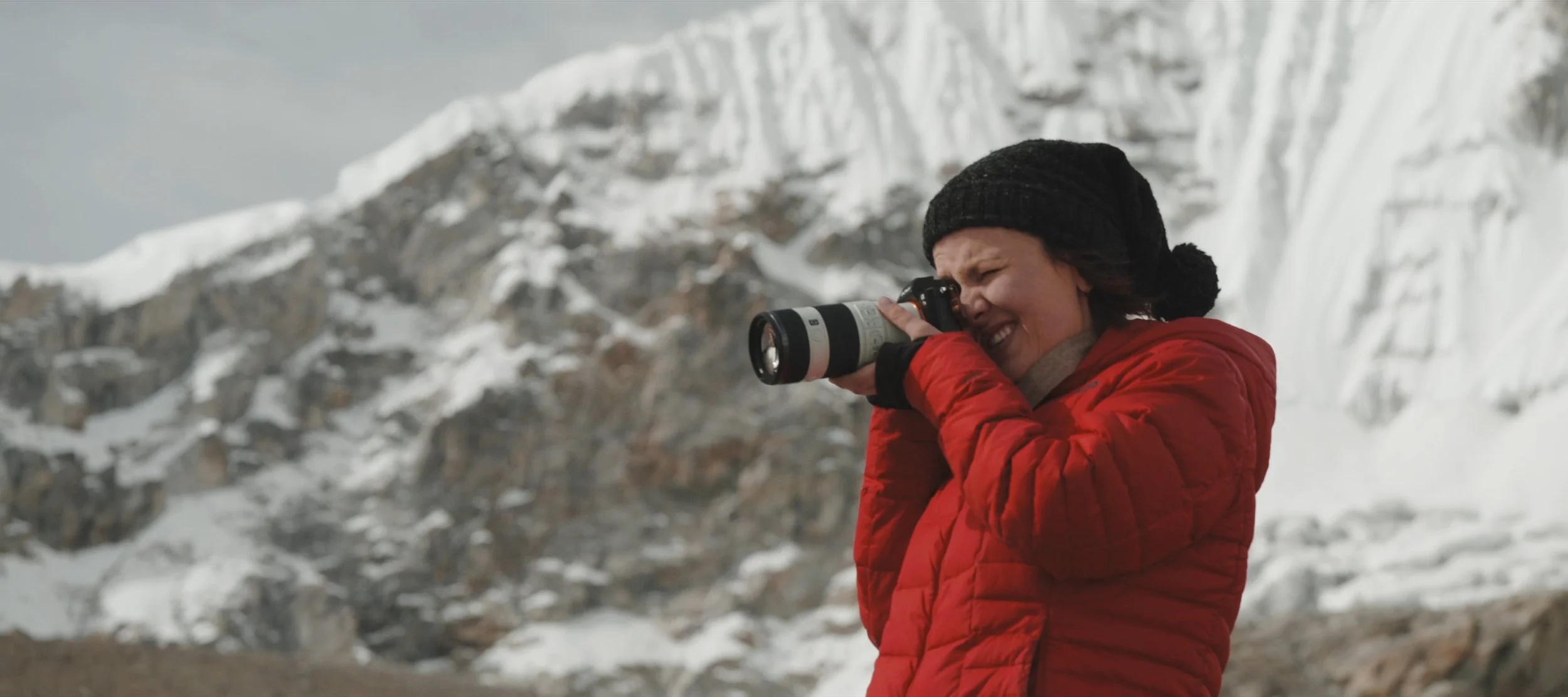 Woman in red jacket and black beanie taking a photo with a camera in snowy mountain landscape