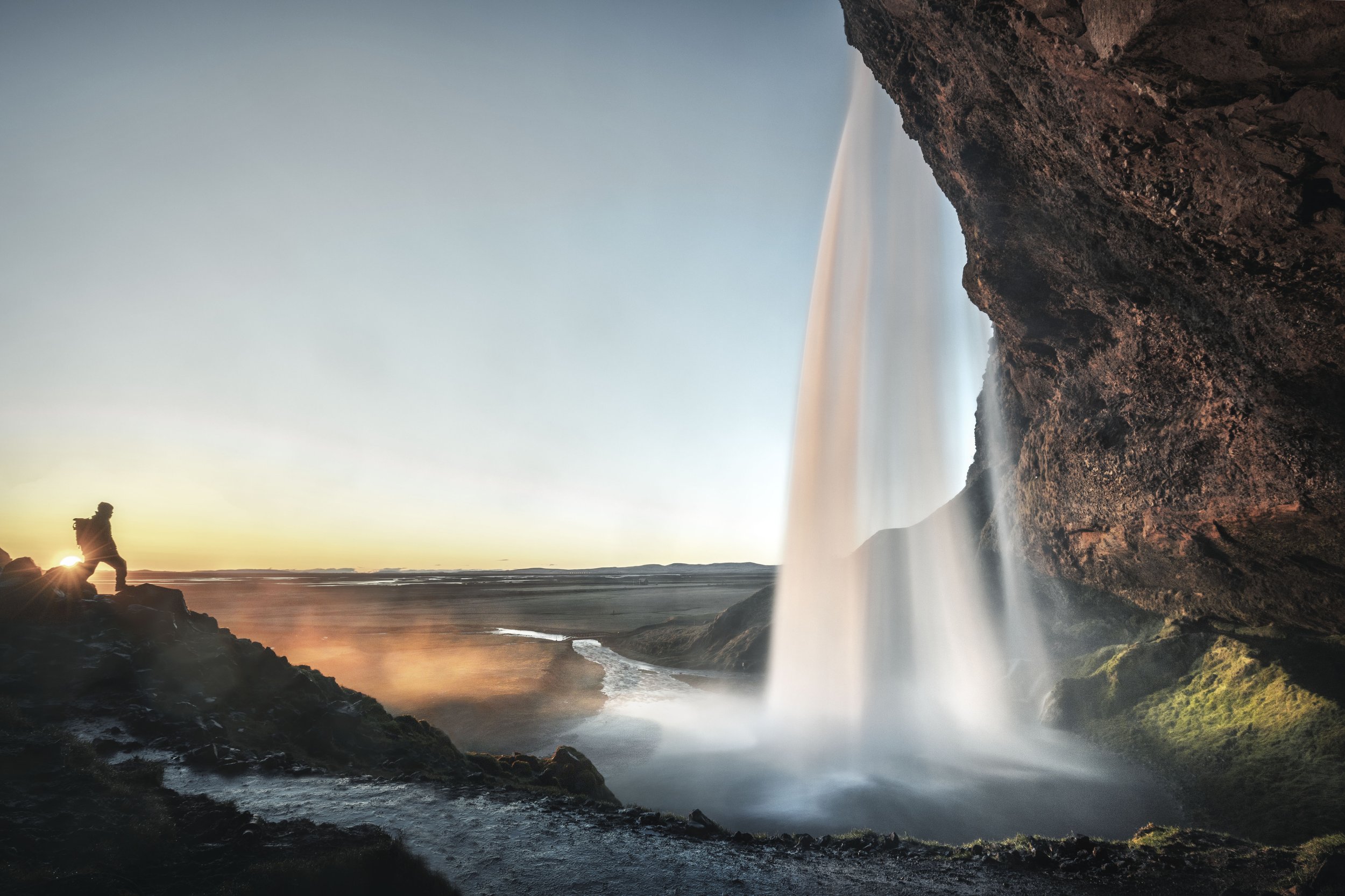 A person standing on rocks near a waterfall at sunset or sunrise, with water flowing over a cliff and a river winding through the landscape.