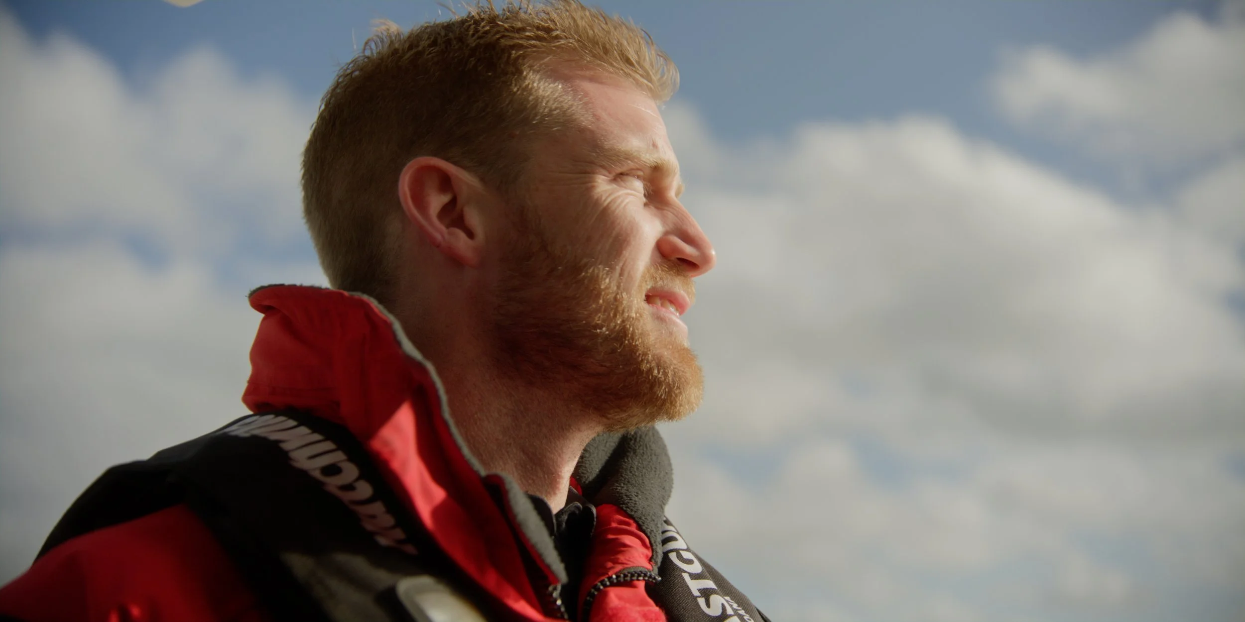 A man with red hair and a beard wearing a red and black jacket looking towards the sky, sunlight on his face, cloudy sky background.