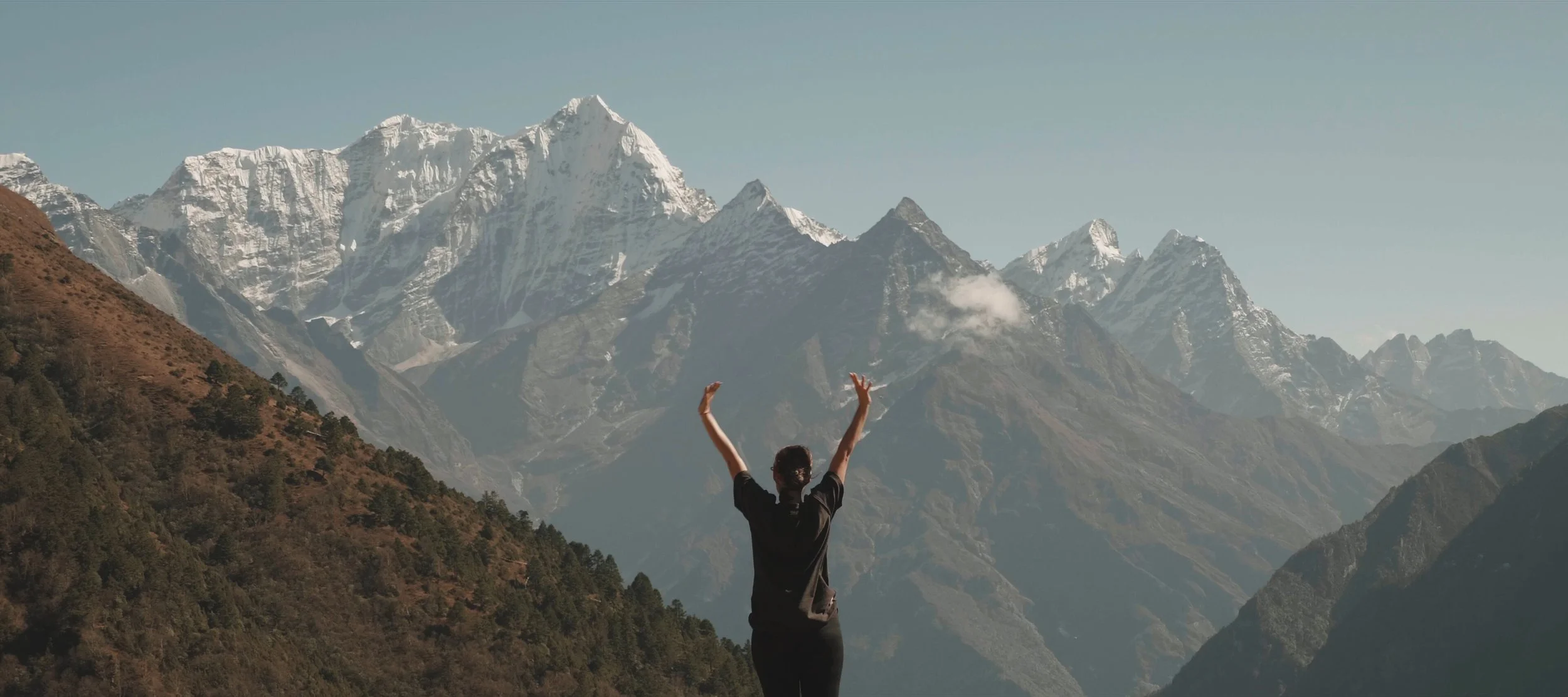 A person standing with arms raised in front of snow-capped mountains and a partly cloudy sky.