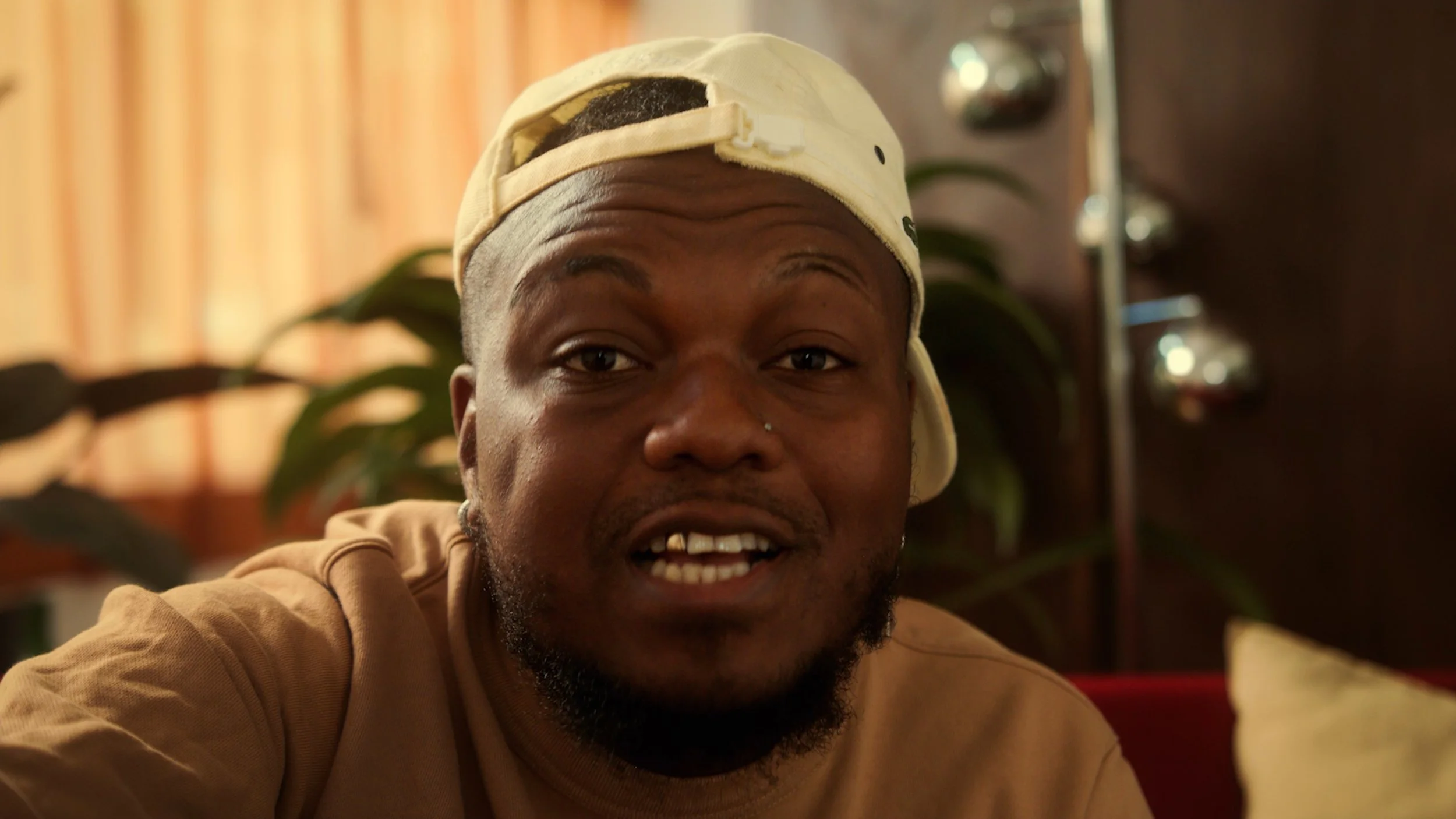 Close-up of a man with brown skin, wearing a beige cap backwards, smiling at the camera, with a background of a plant and a lamp.