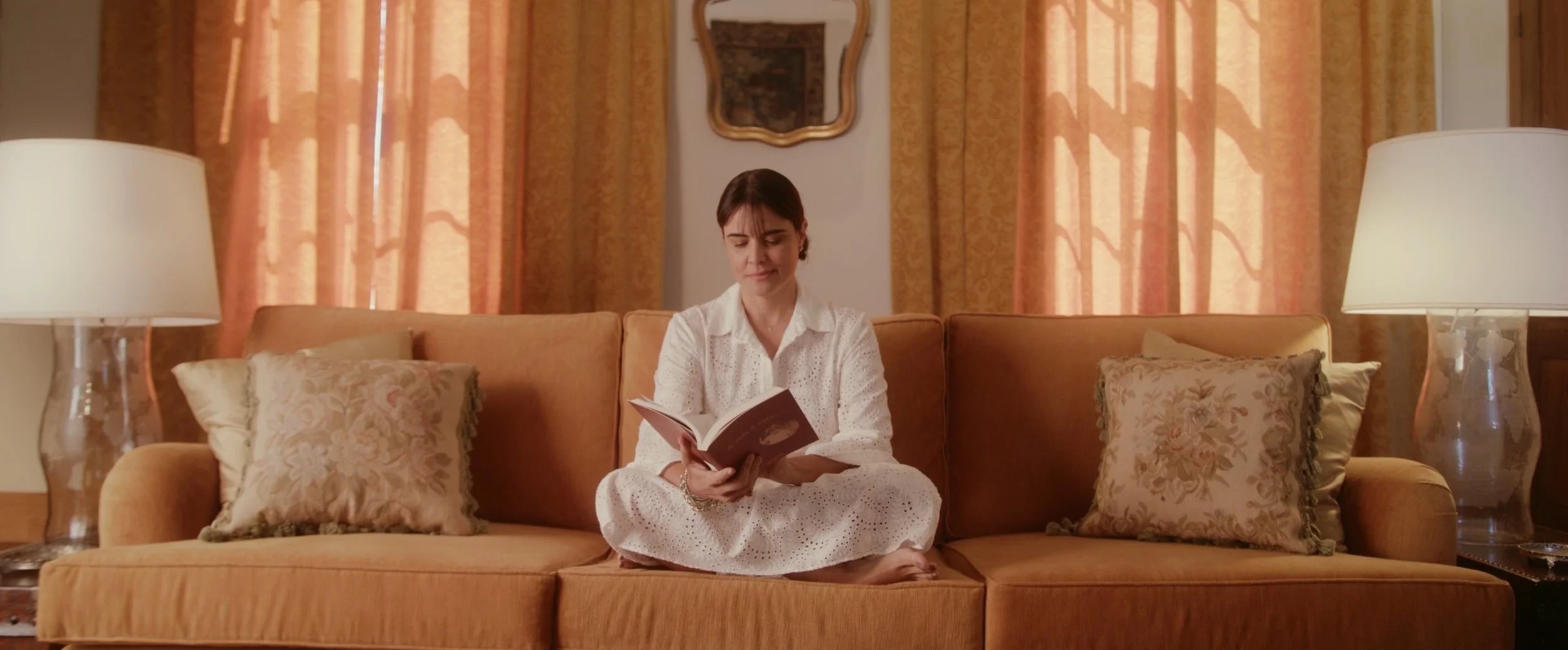 Woman sitting cross-legged on an orange sofa, reading a book, surrounded by lamps and patterned curtains.