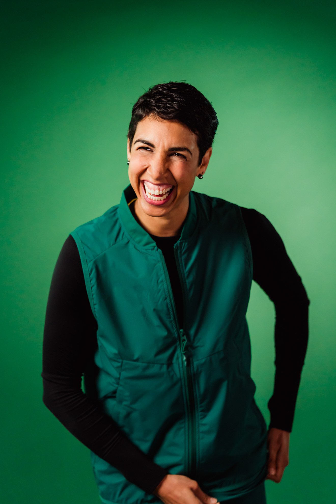 A woman with short dark hair wearing a black long sleeve shirt and teal vest, smiling broadly against a green background.