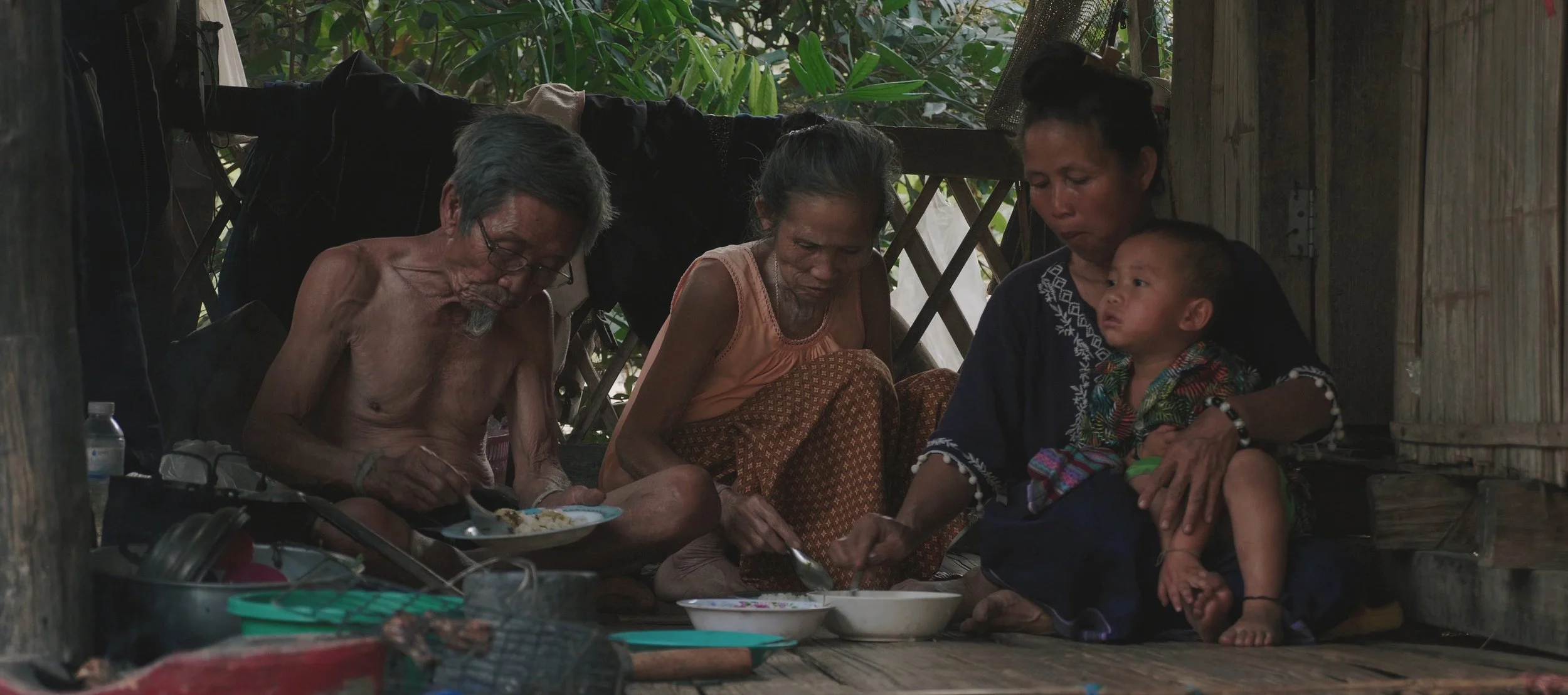 Family sitting on wooden floor inside a rustic home, sharing a meal together.