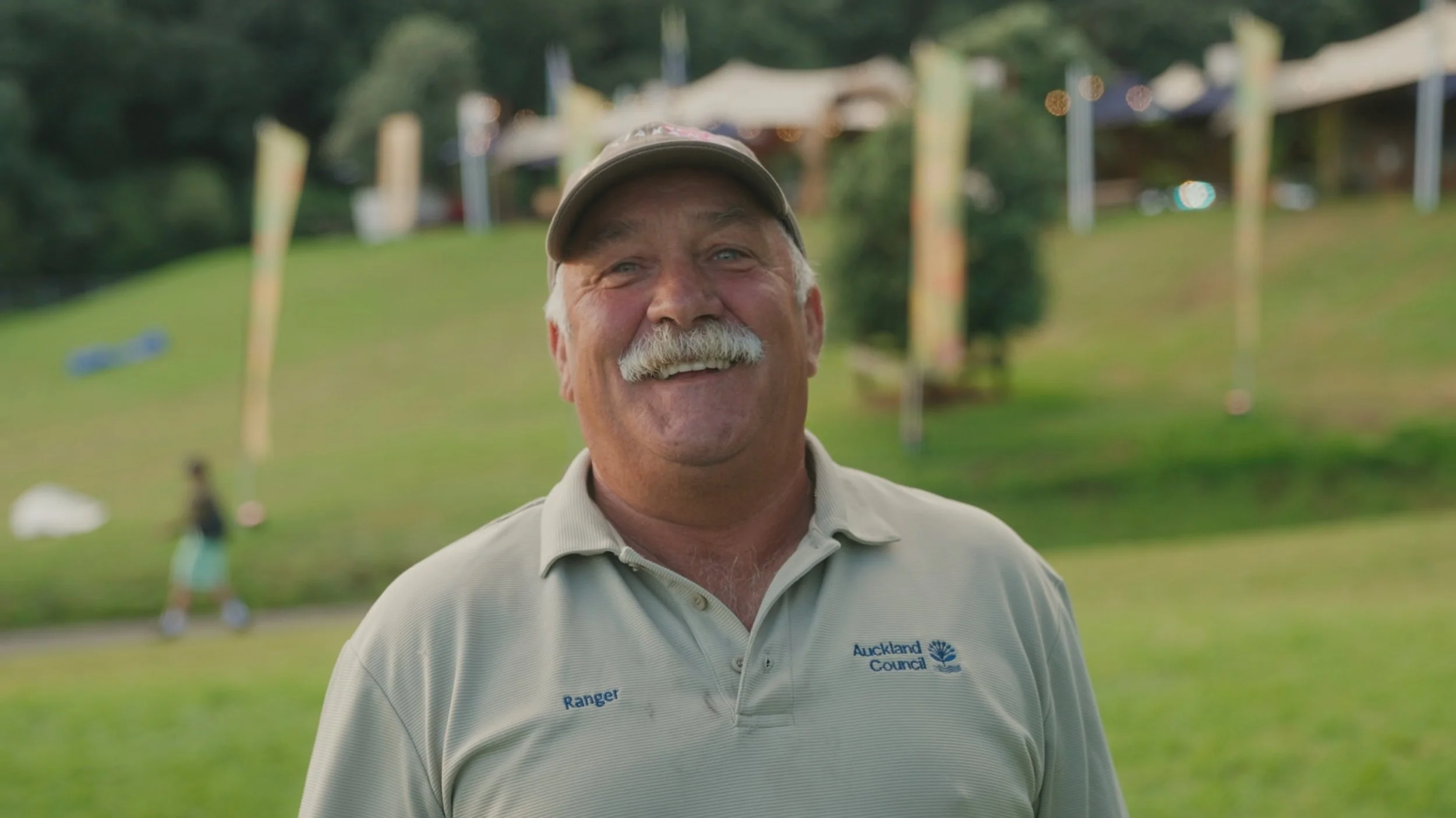 A smiling man with a mustache and gray hair wearing a beige shirt and cap, standing outdoors in a grassy area with trees and a playground in the background.