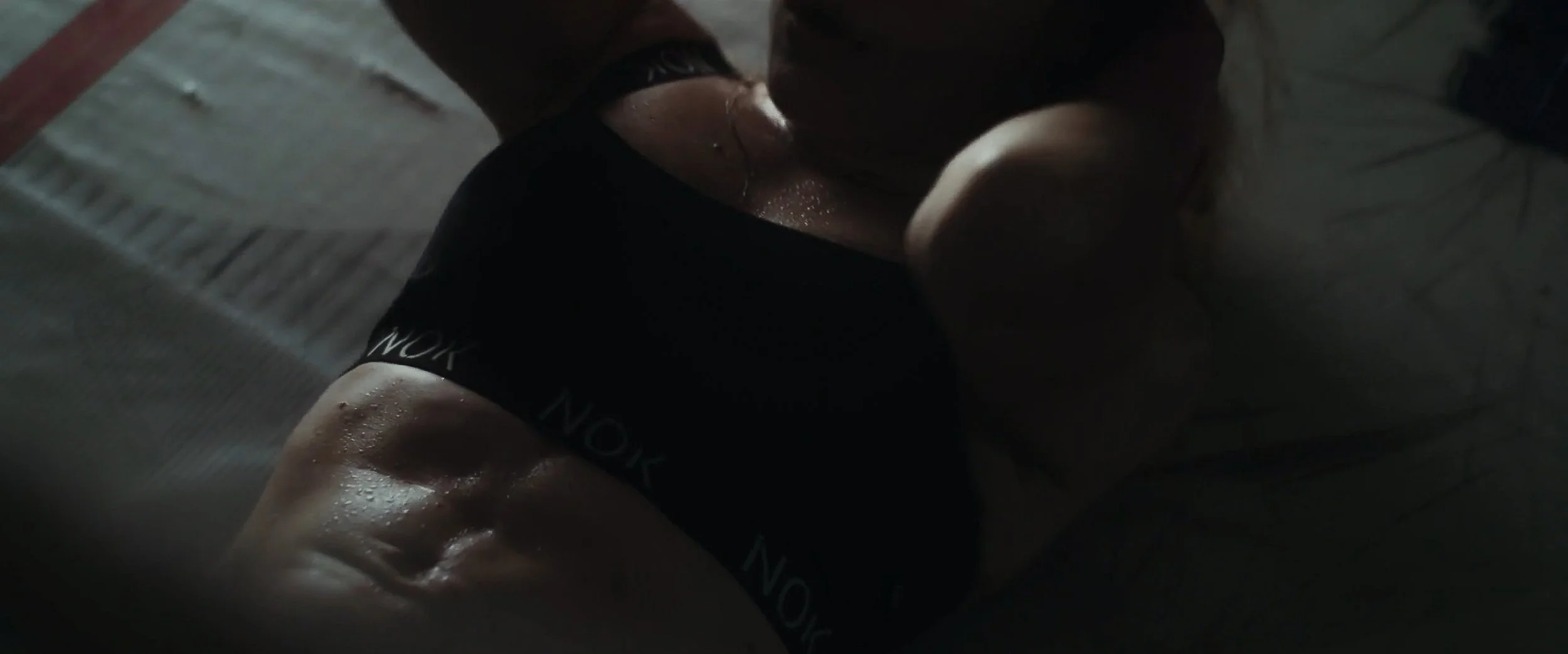 Person lying on a hard floor, wearing black shorts with white text, wet with sweat, in a dimly lit environment.