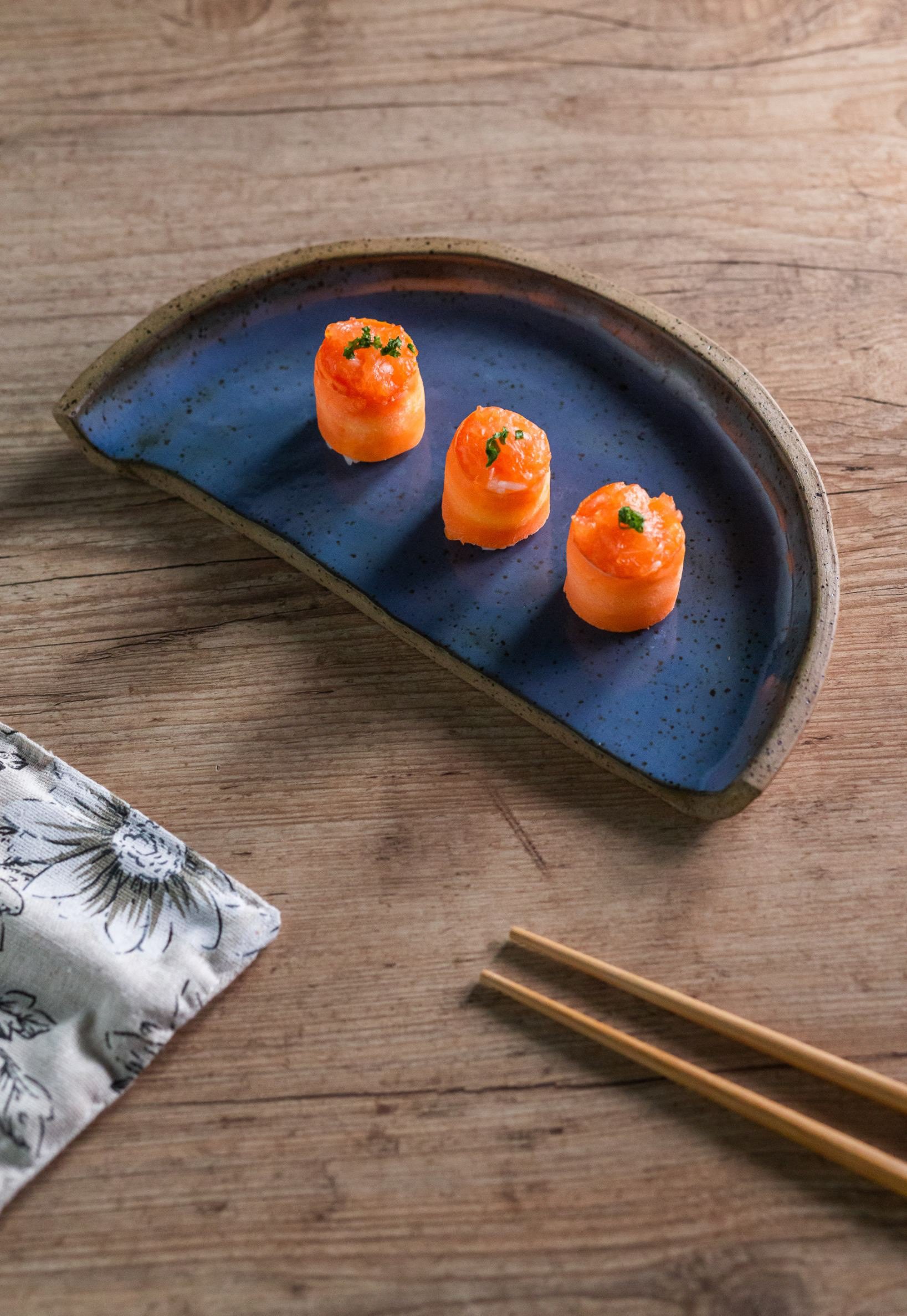Three pieces of sushi wrapped in salmon with green garnish on a blue ceramic plate, on a wooden table with chopsticks and a floral cloth napkin nearby.