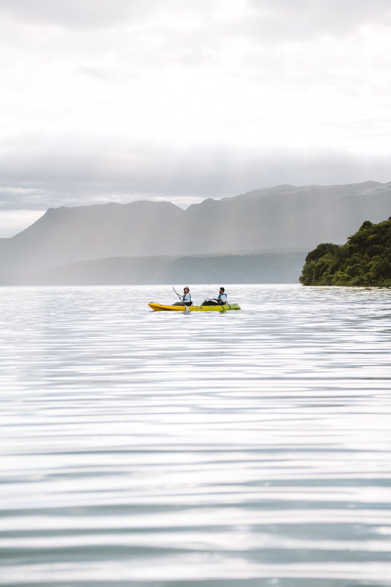 Two people in a yellow kayak on a calm lake with mountains and cloudy sky in the background.