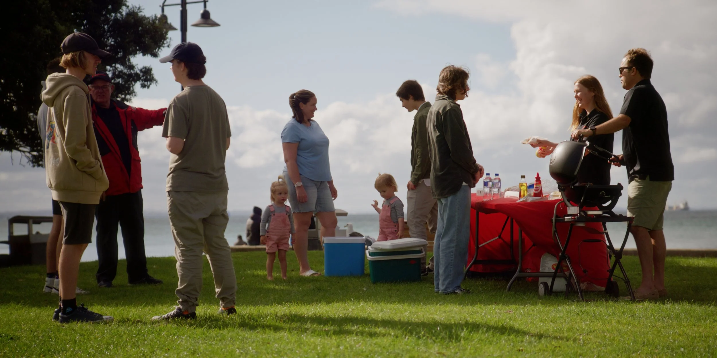 Group of people gathered outdoors near a red table with food and drinks, preparing for a barbecue by the water on a cloudy day.