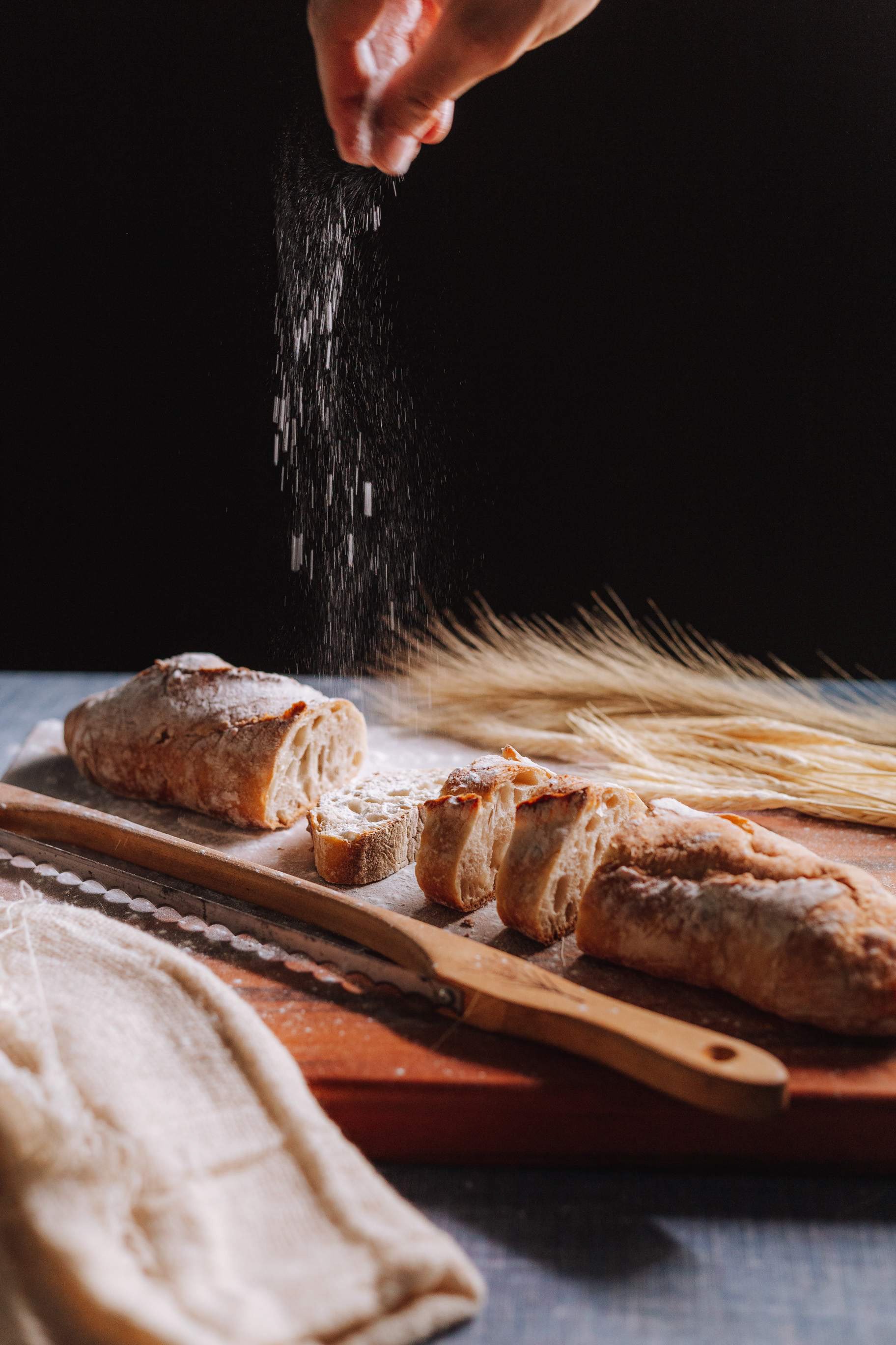 Hand sprinkling flour on freshly baked bread on a cutting board with pasta and a cloth nearby