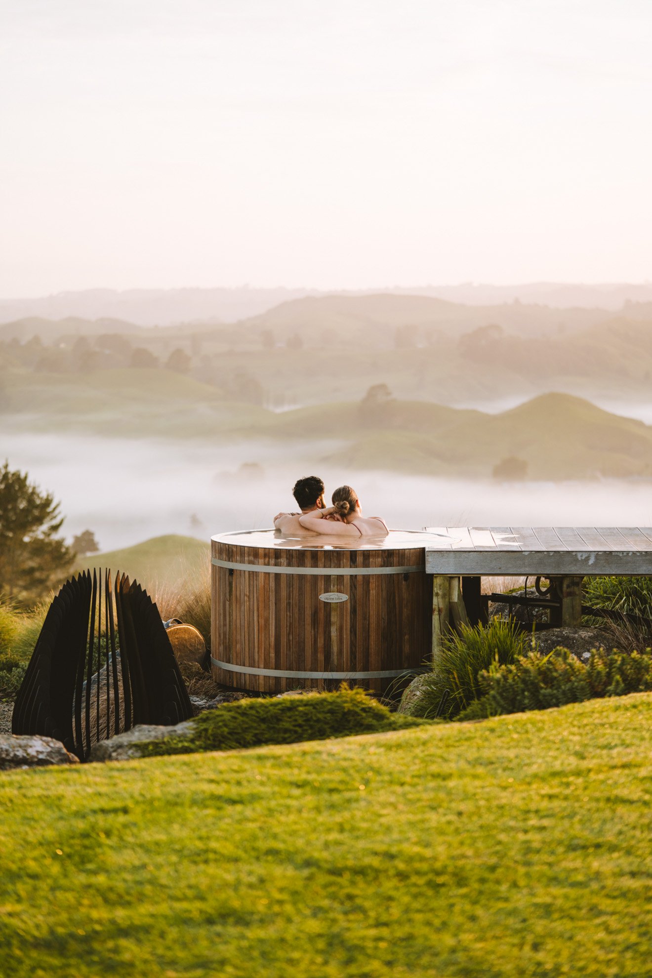 Couple relaxing in a hot tub outdoors with scenic rolling hills and fog in the background.
