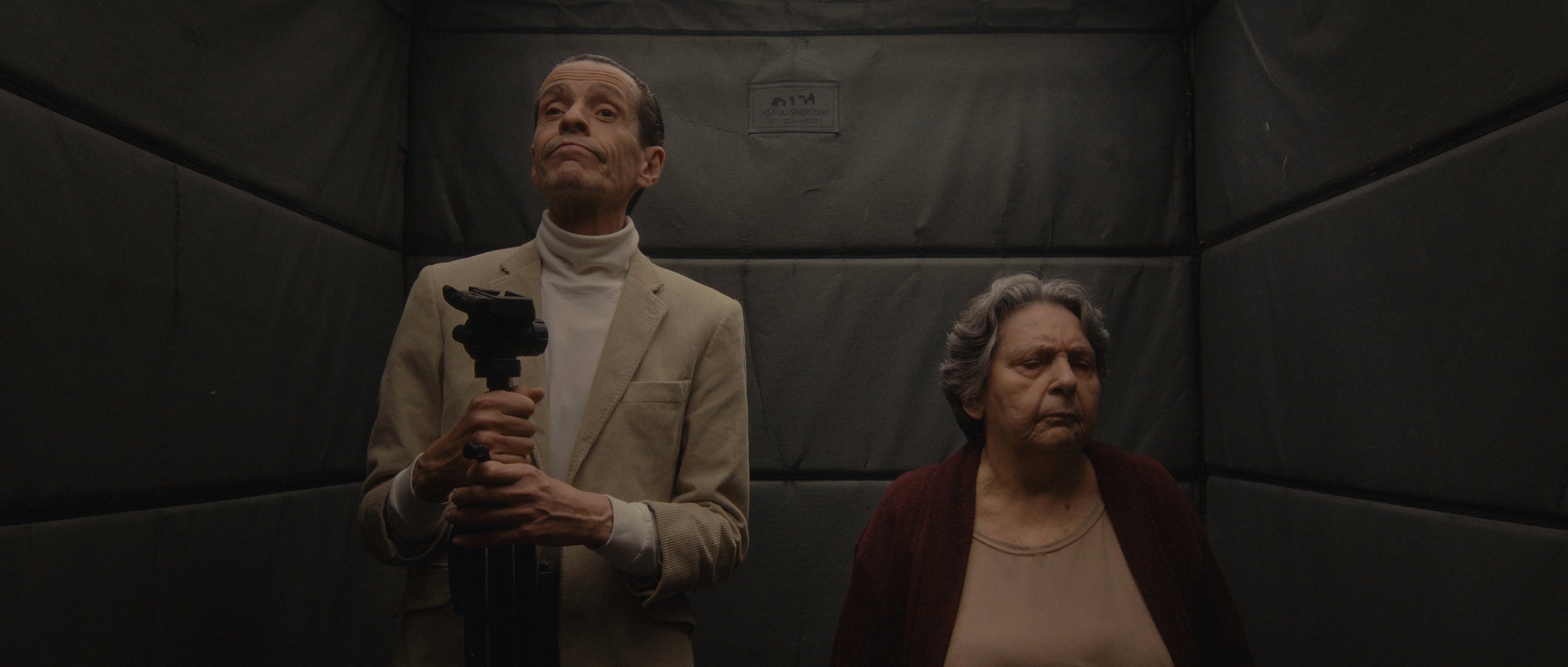 A man in a beige blazer and white turtleneck standing with a serious expression, holding a gun-shaped object, in an elevator with a woman in a dark cardigan standing next to him, looking down with a serious expression.
