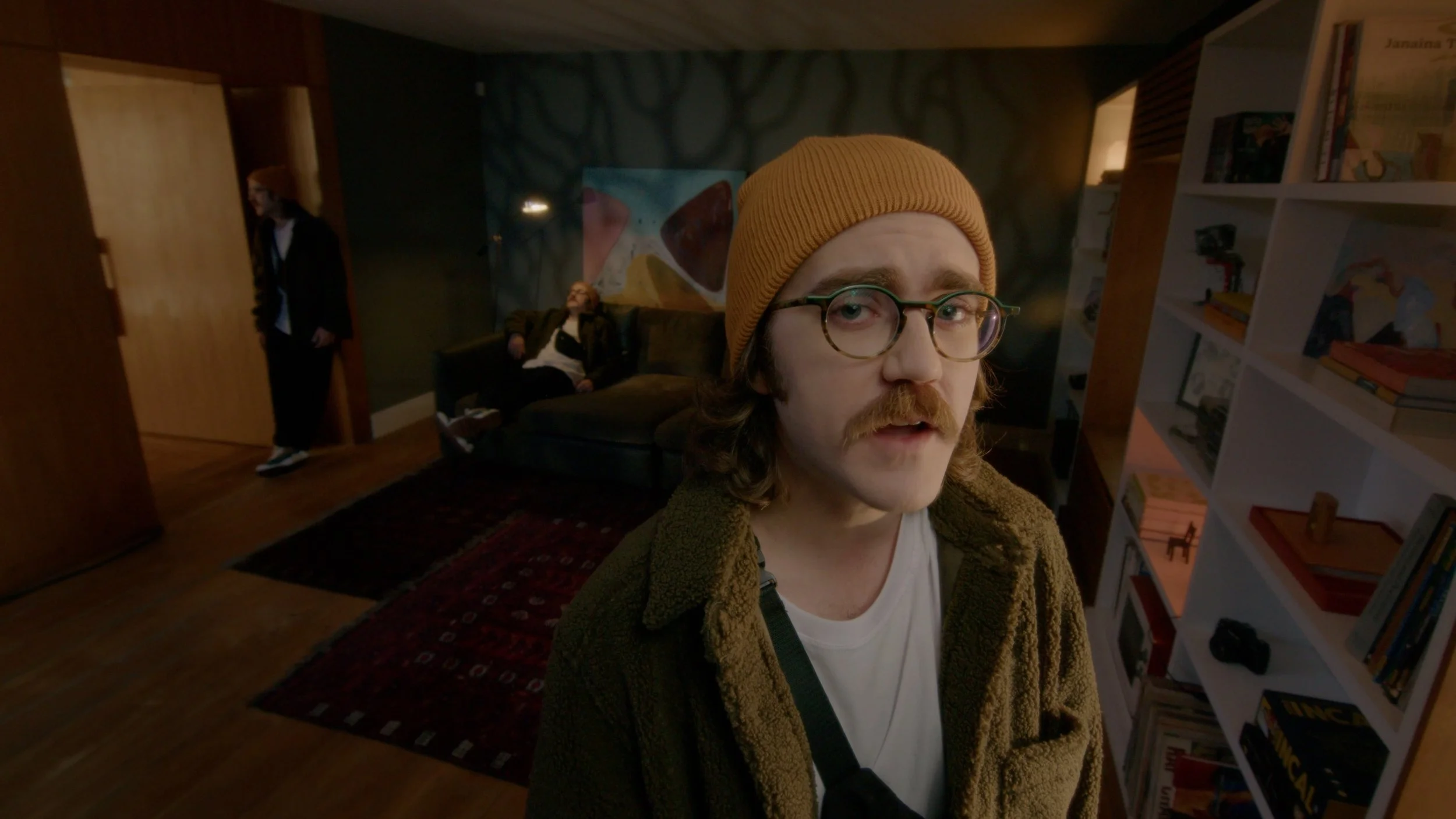 A young man with glasses, a mustache, and long hair, wearing a yellow beanie and brown jacket, looks into the camera inside a room with bookshelves. Two other people are in the background, one sitting on a couch and the other standing near a doorway.